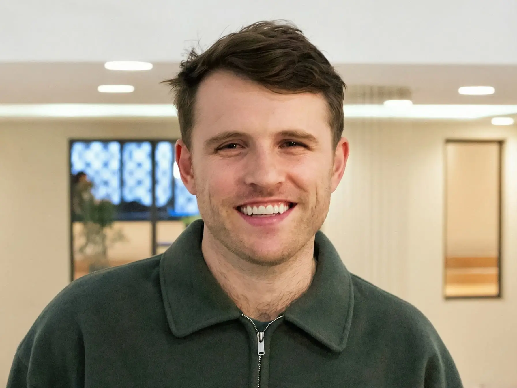 Smiling man in a dark green zip-collar fleece stands in a softly lit indoor room.