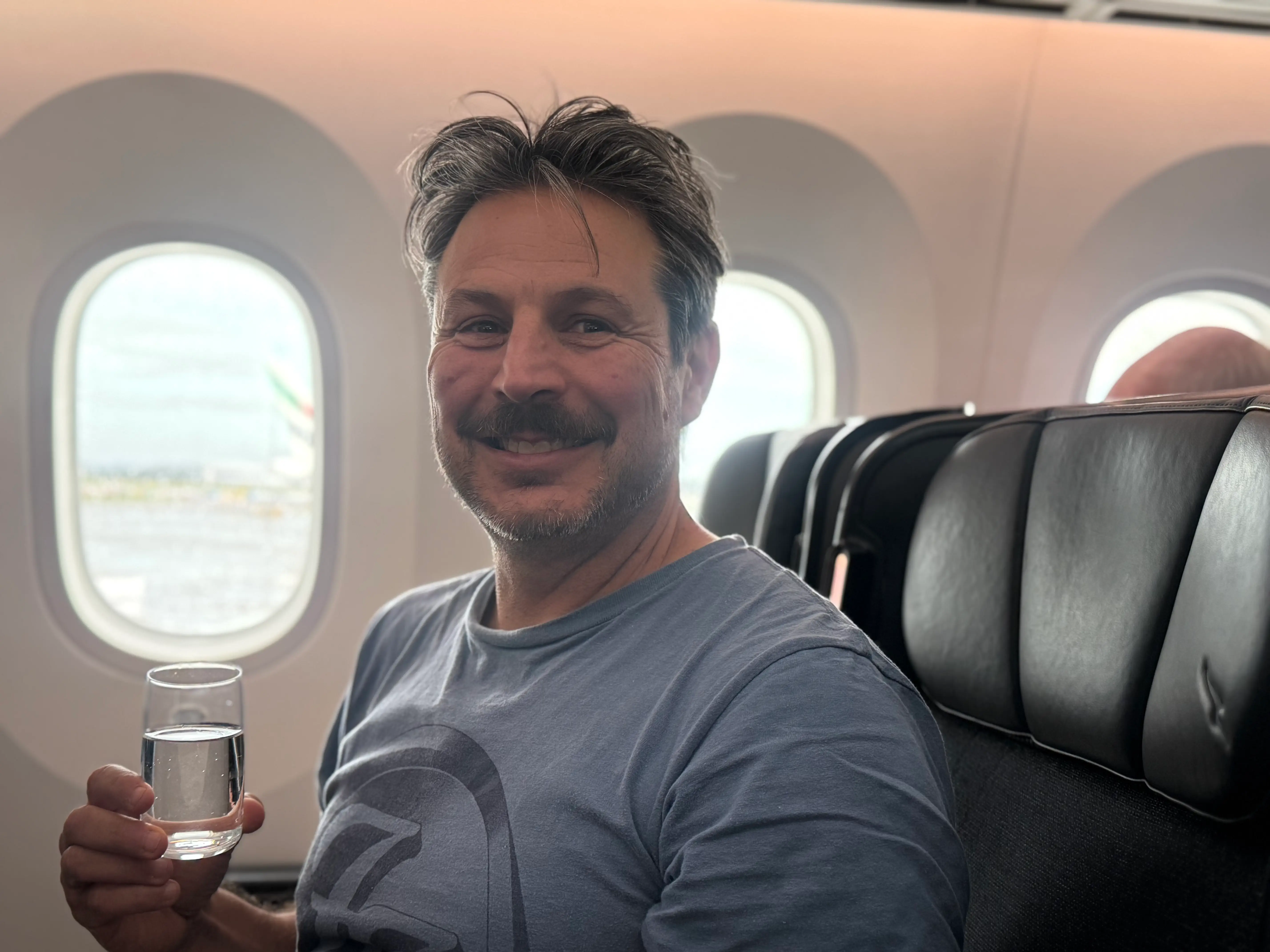 Author Ash Jurberg in premium economy seat with drink, smiling