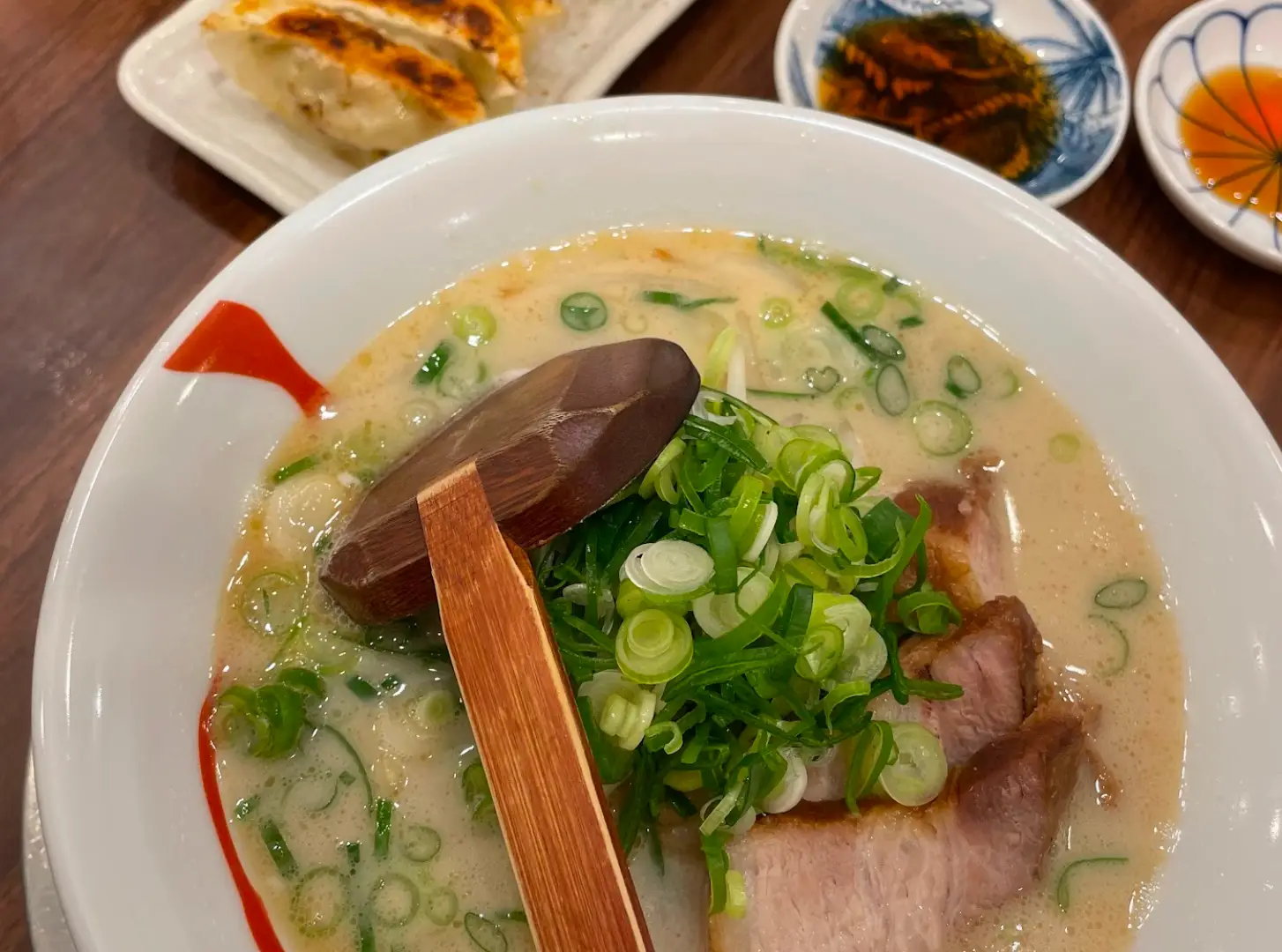 Bowl of creamy ramen topped with sliced pork and green onions, with gyoza and dipping sauces on a table.