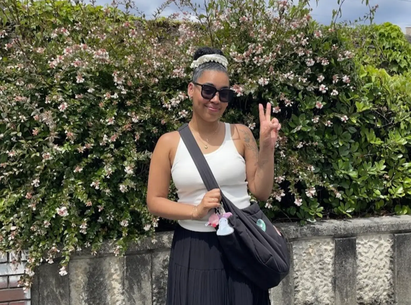 The writer wearing a white top and black skirt and holding up a peace sign in front of a flowering plant.
