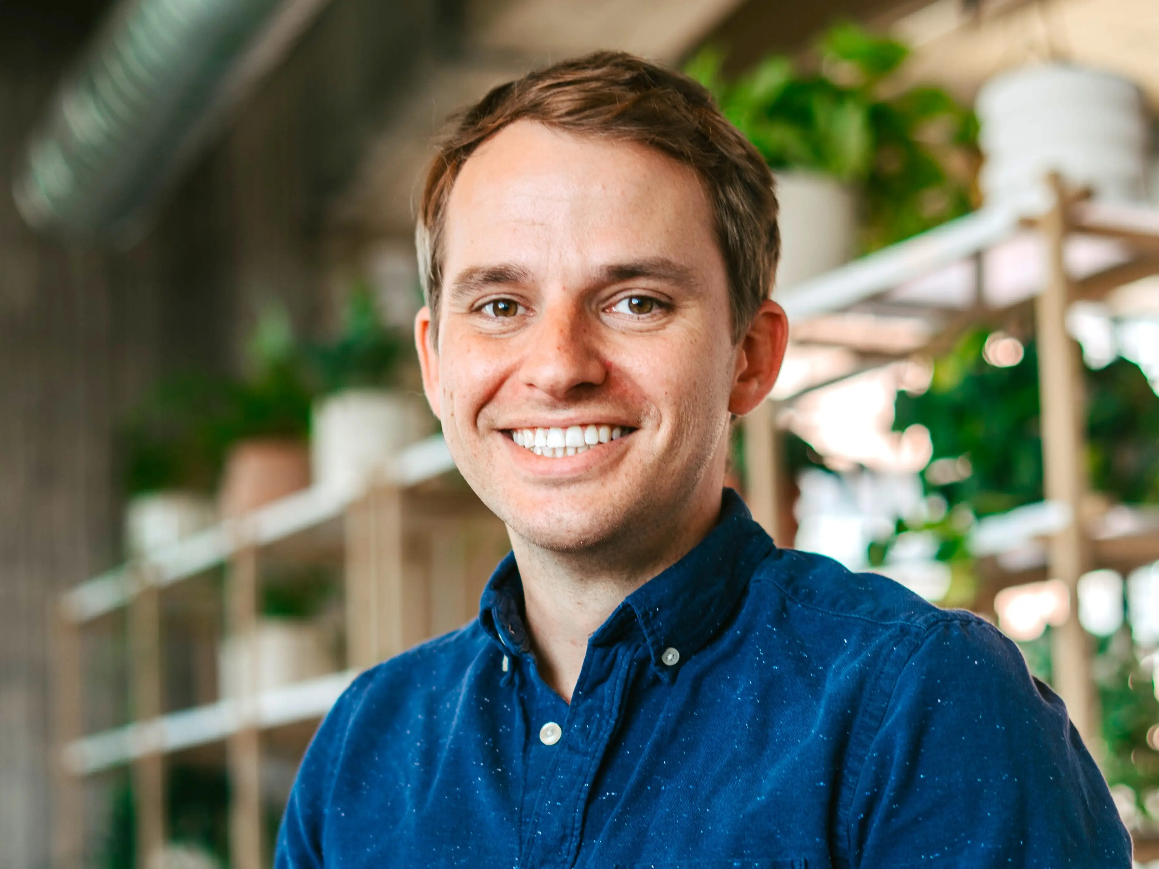 Smiling man in a blue button-up shirt seated indoors beside shelves with potted plants.