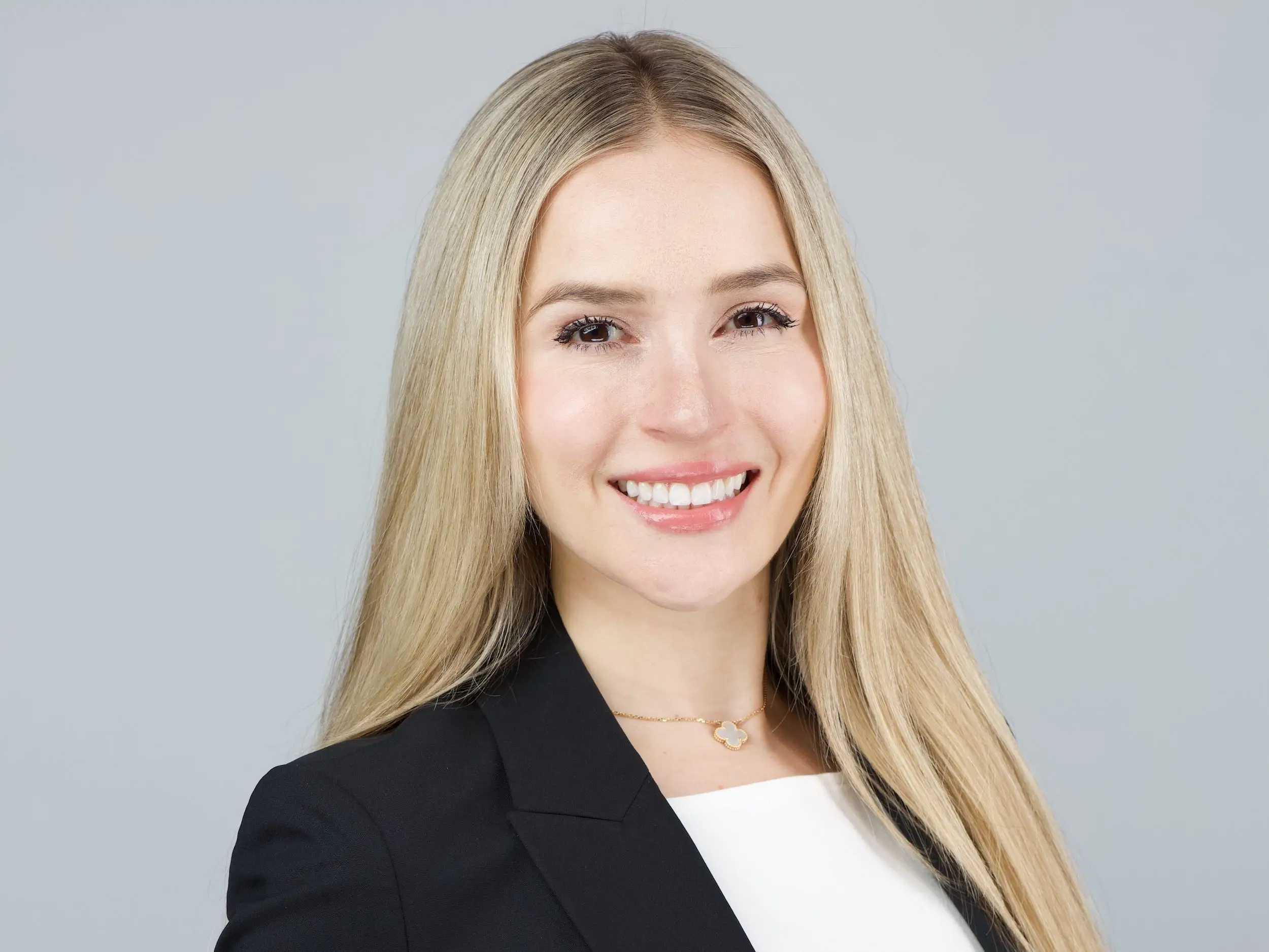 Smiling woman with long blond hair poses in a black blazer against a plain gray studio background.