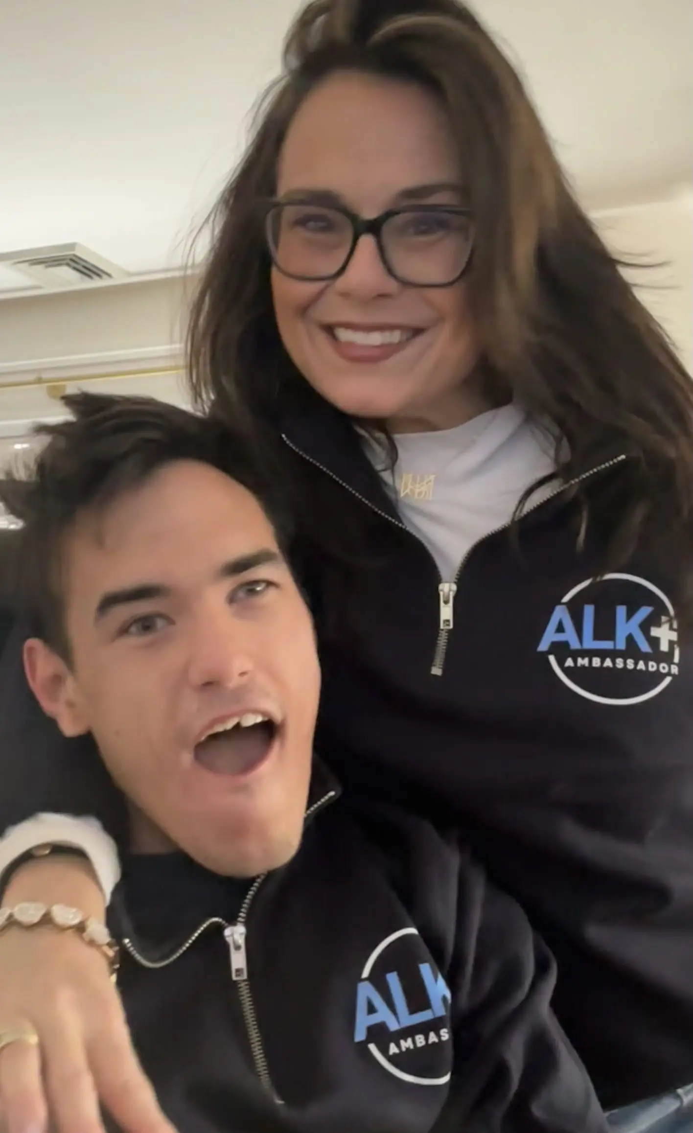 Two smiling people pose together indoors wearing black zip-up tops with ALK+ AMBASSADOR logos.