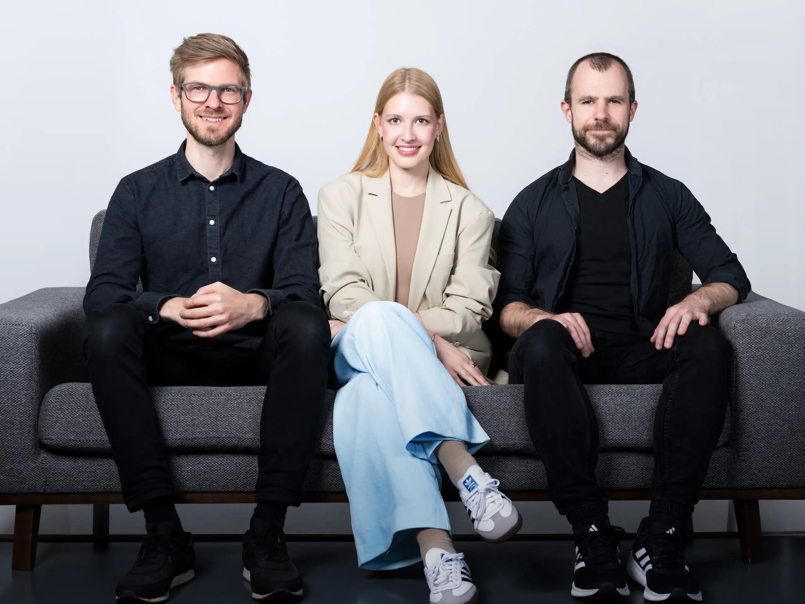 Three people sit together on a gray sofa against a plain studio background.