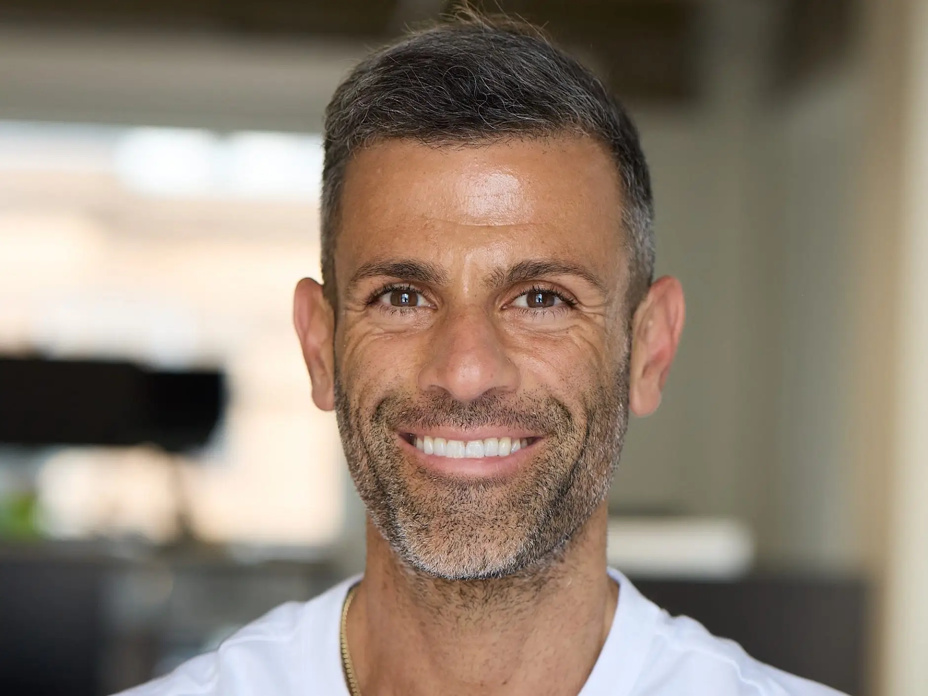 Smiling man in a white T-shirt poses with arms crossed in a softly lit indoor setting.