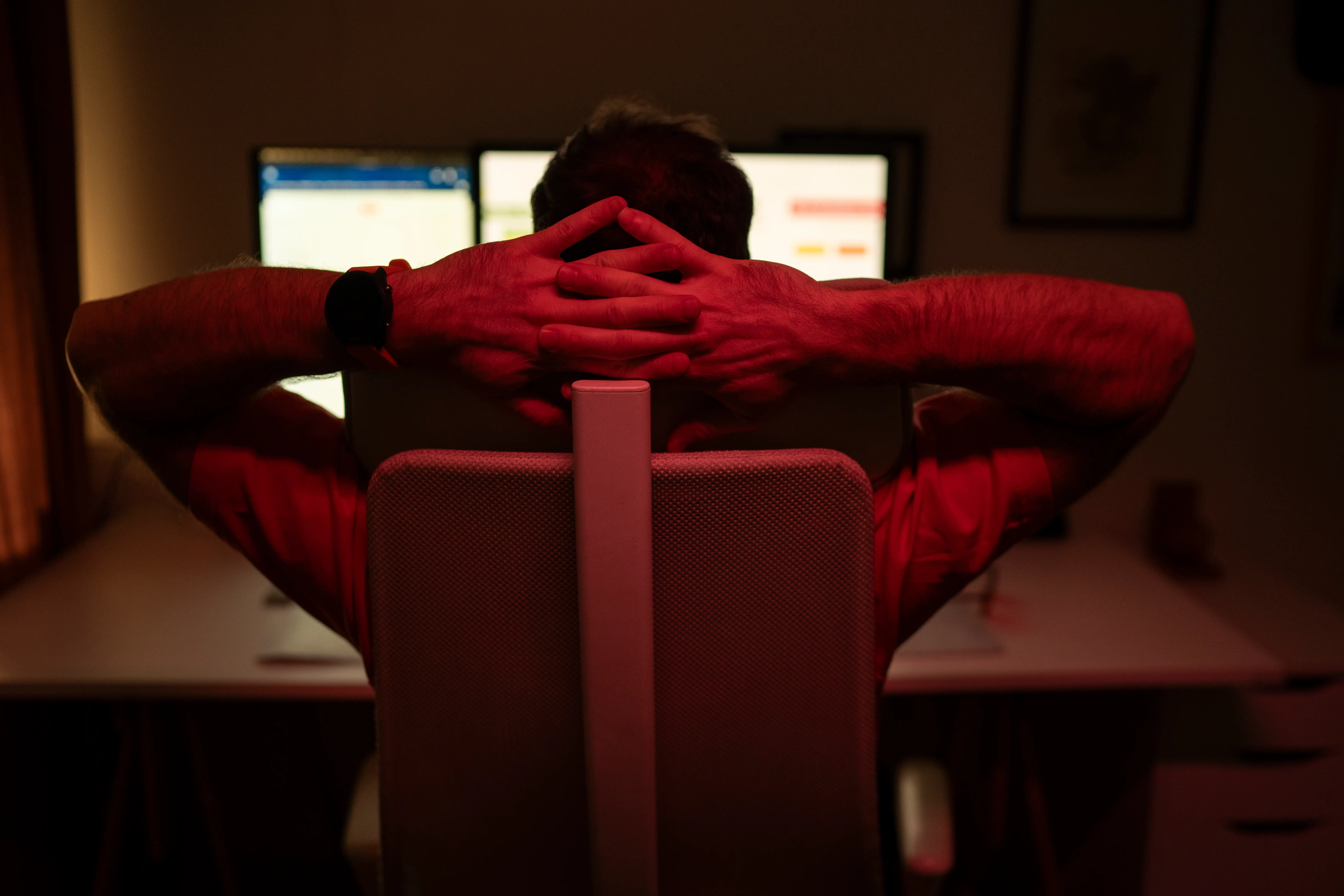 Person seated at a desk with hands clasped behind head facing two computer monitors in dim red lighting.