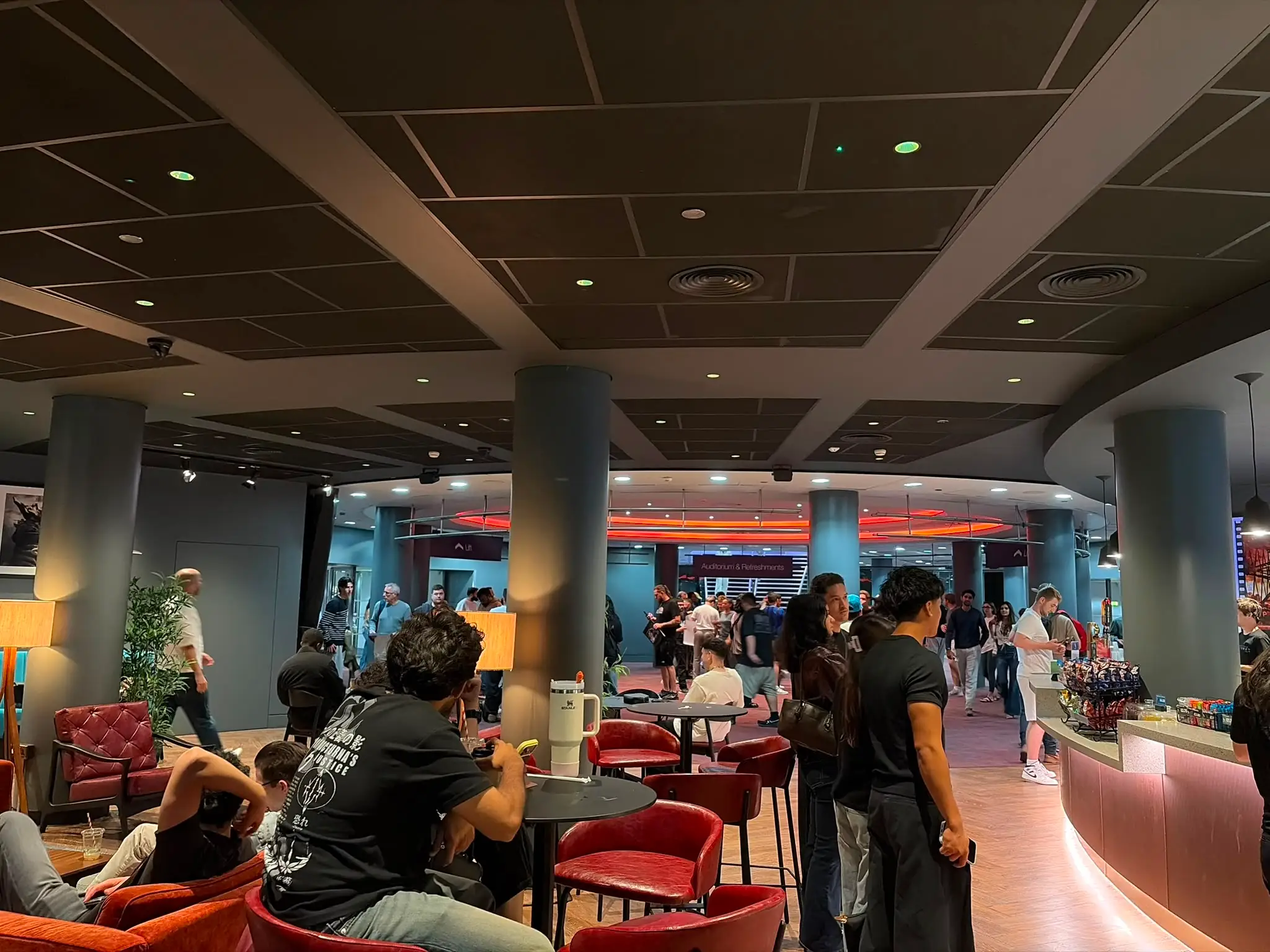 Crowded indoor lounge with red seating, round tables, columns, and a lit service counter in the background.