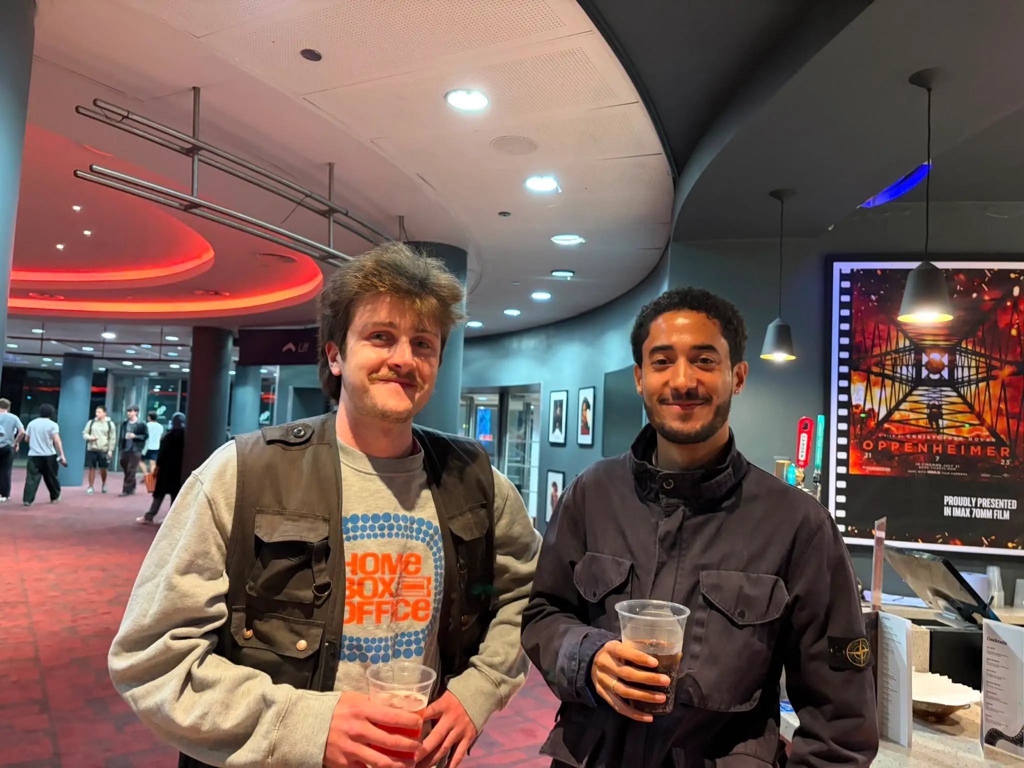 Two people holding drinks stand in a cinema lobby beside an OPPENHEIMER IMAX 70MM FILM poster.