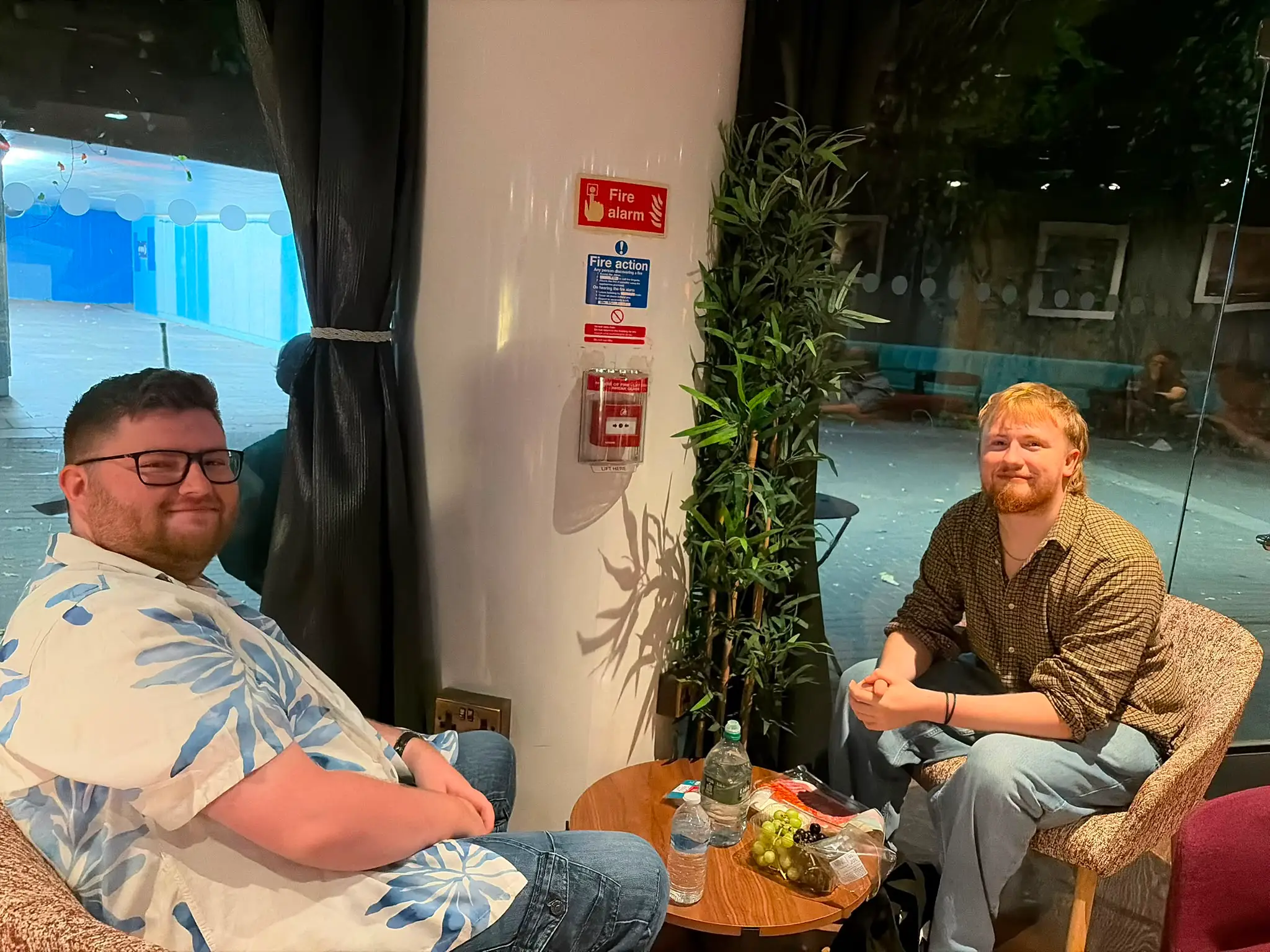 Two people sit by a small table in an indoor lounge beside a window and fire alarm signs.