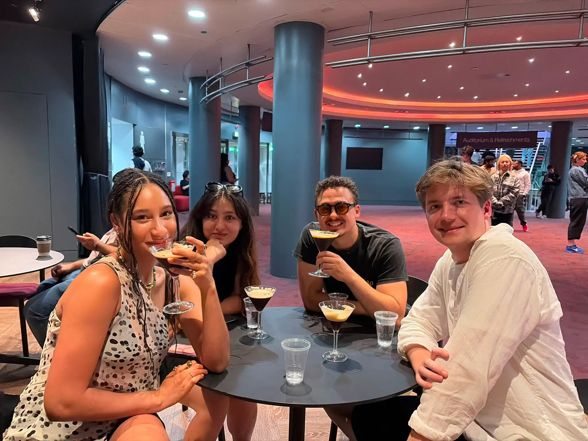 Four people sit around a table with cocktails in a modern auditorium lobby.