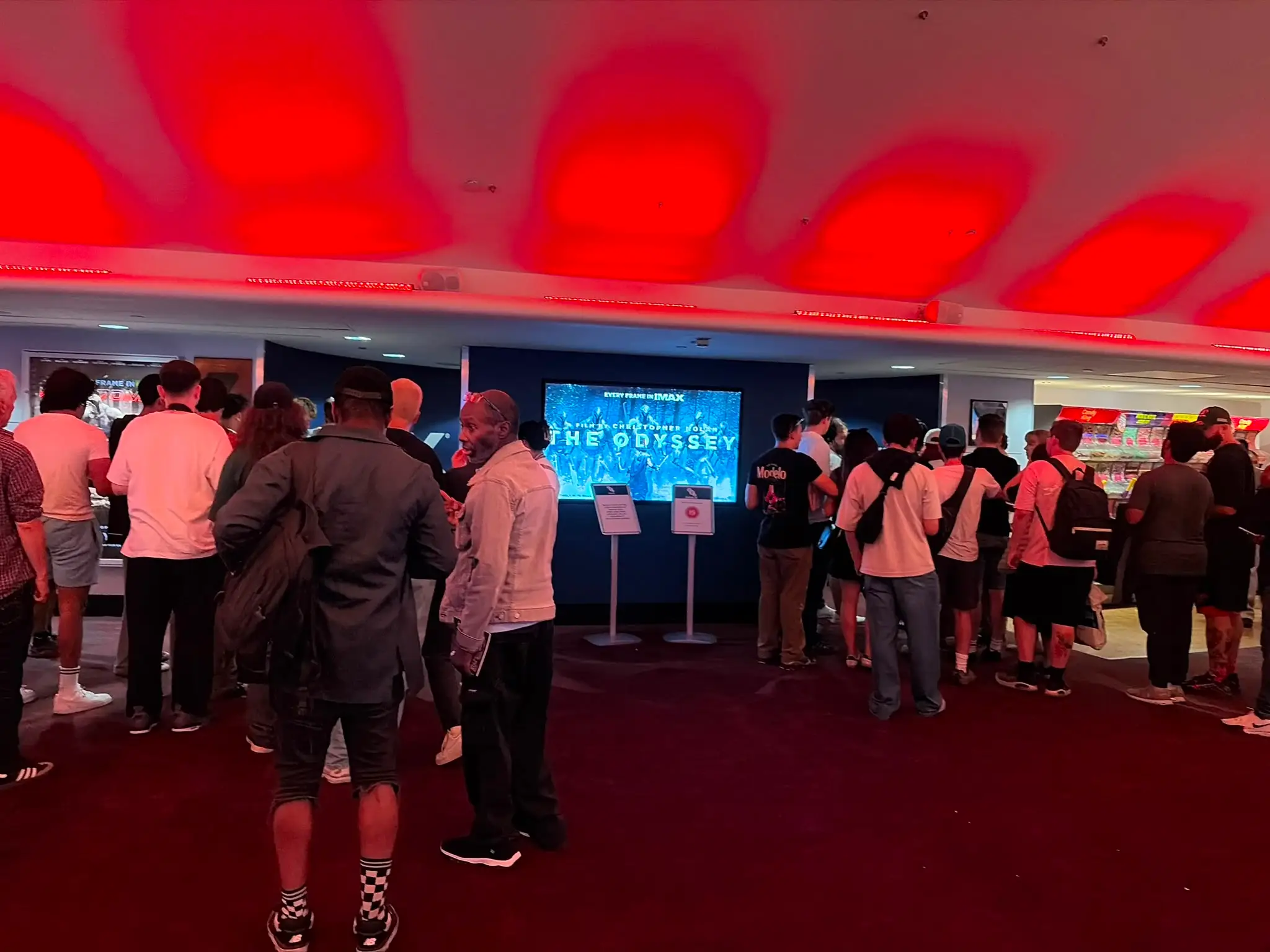 Crowds queue in a red-lit cinema lobby beside a digital display for THE ODYSSEY.