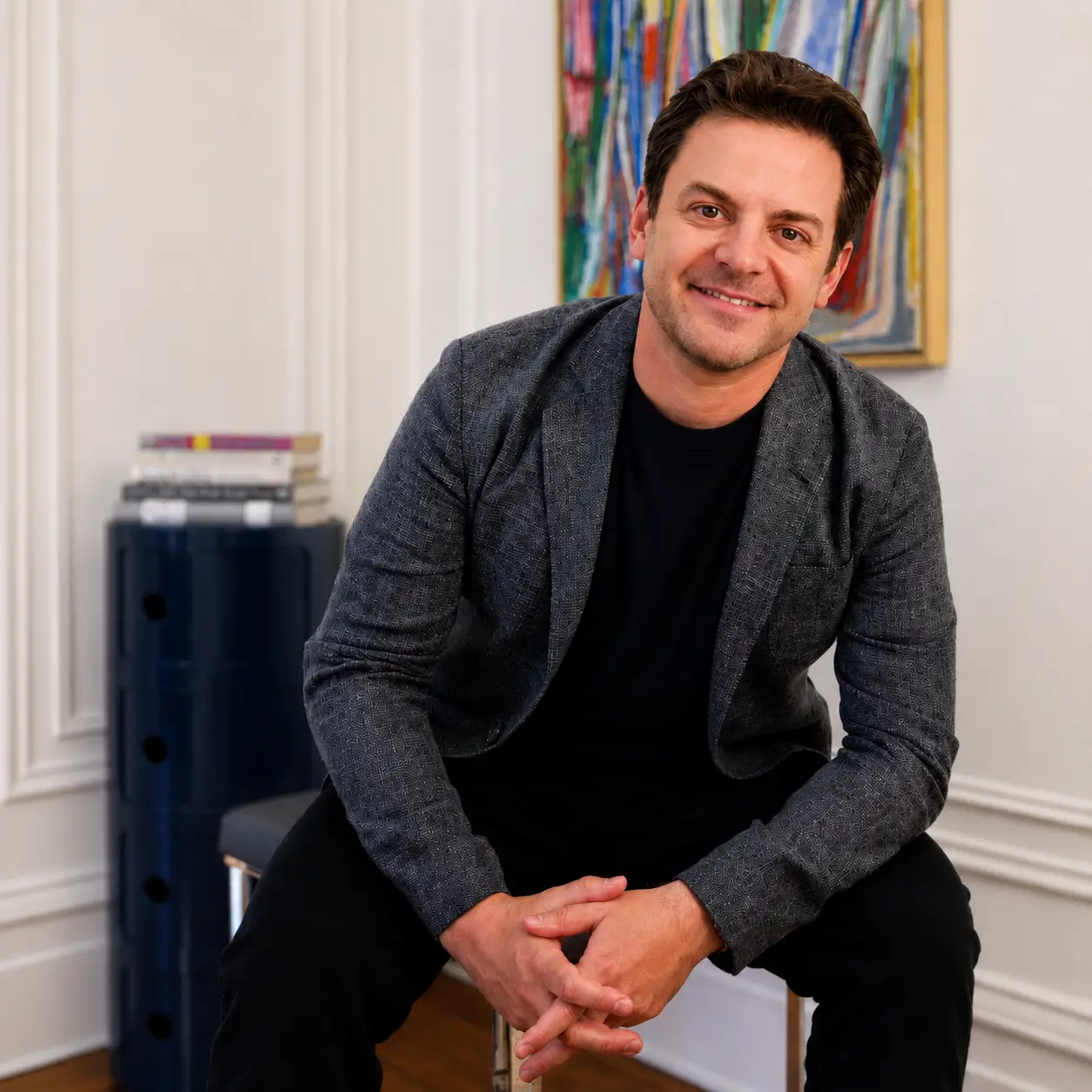 A man in a gray blazer, black shirt, and black pants, sitting on a chair and smiling at the camera.
