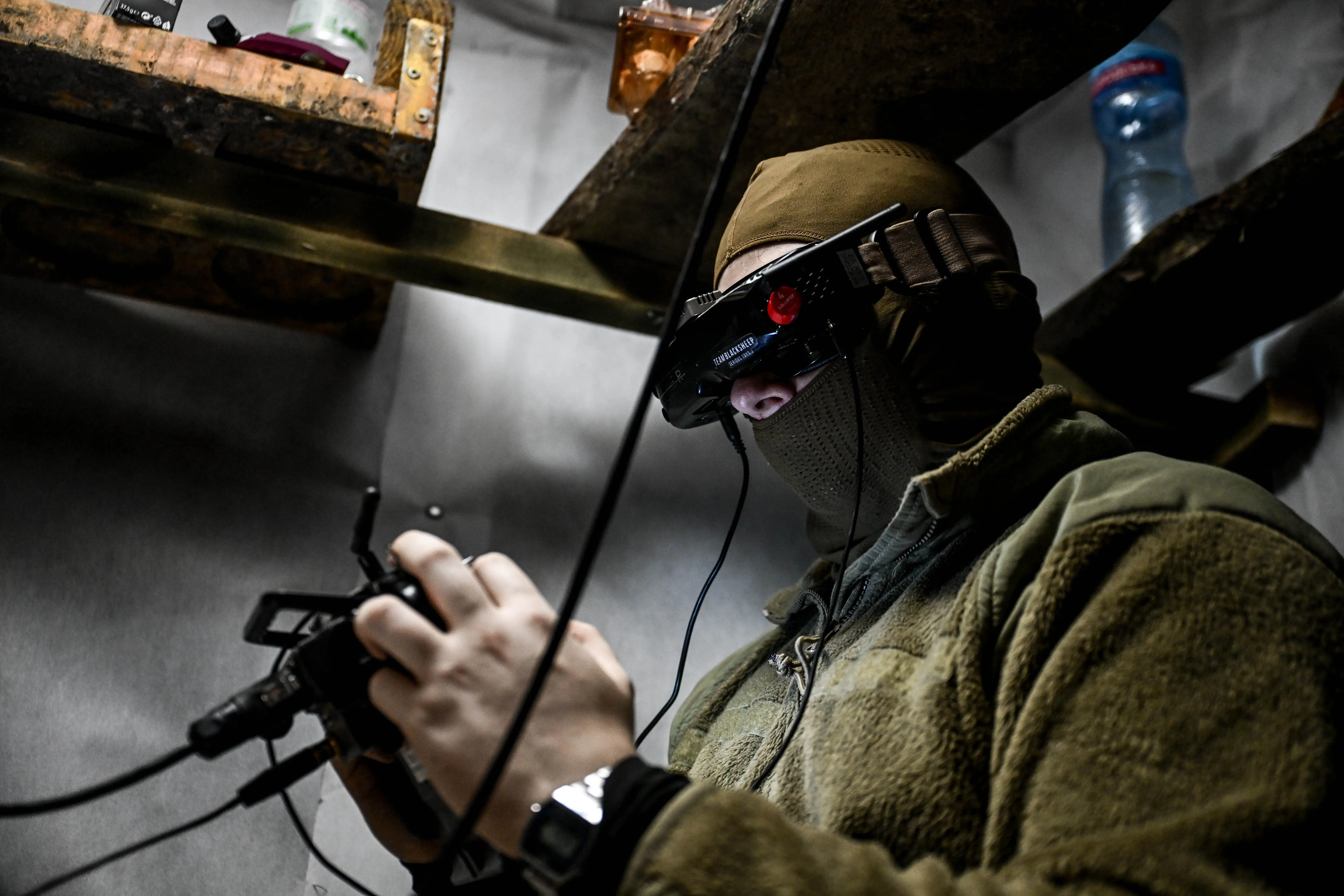 Masked person in tactical-style clothing operates a wired handheld controller while wearing electronic goggles indoors.