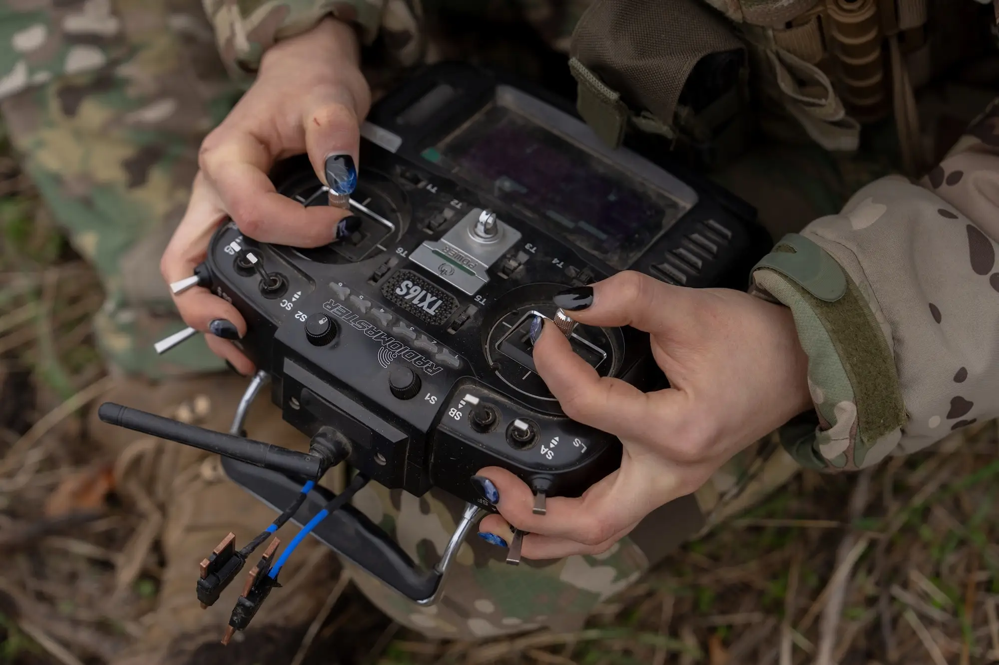 Hands in camouflage clothing operate a RadioMaster TX16S remote controller outdoors among grass and leaves.