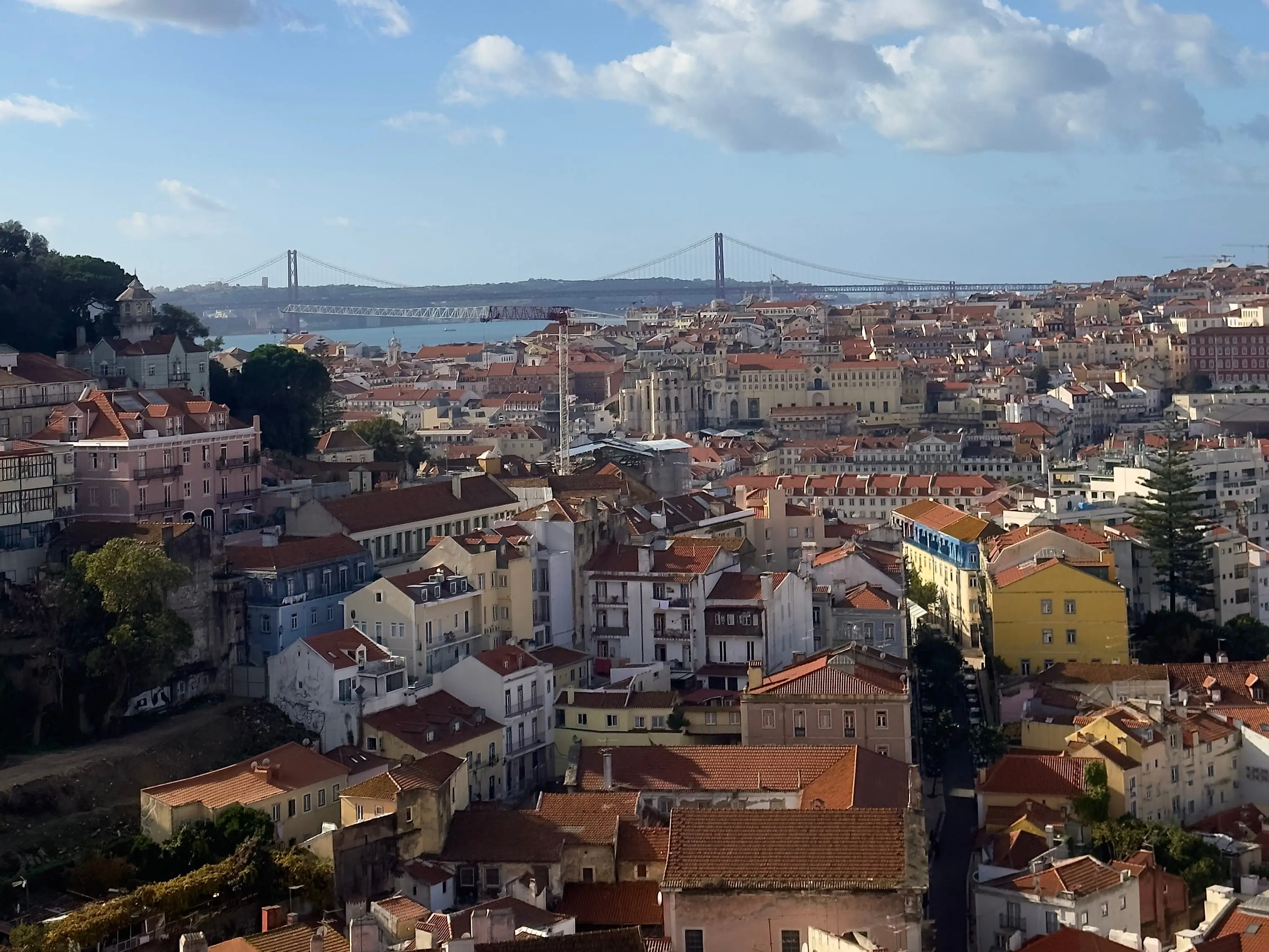 Lisbon rooftops stretch toward the 25 de Abril Bridge under a partly cloudy blue sky.