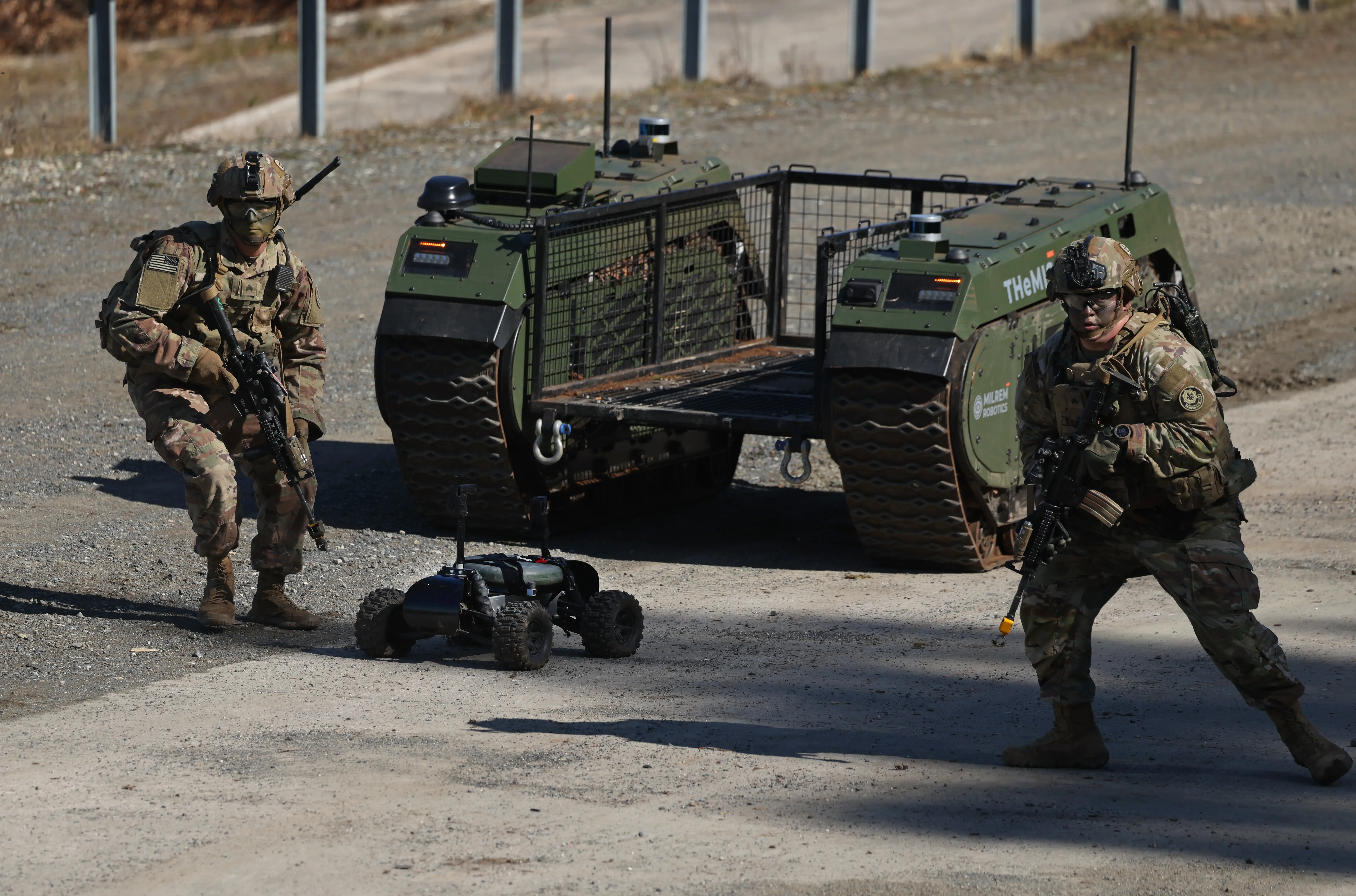 Military personnel maneuver beside an unmanned ground vehicle and a small robot on a gravel road.