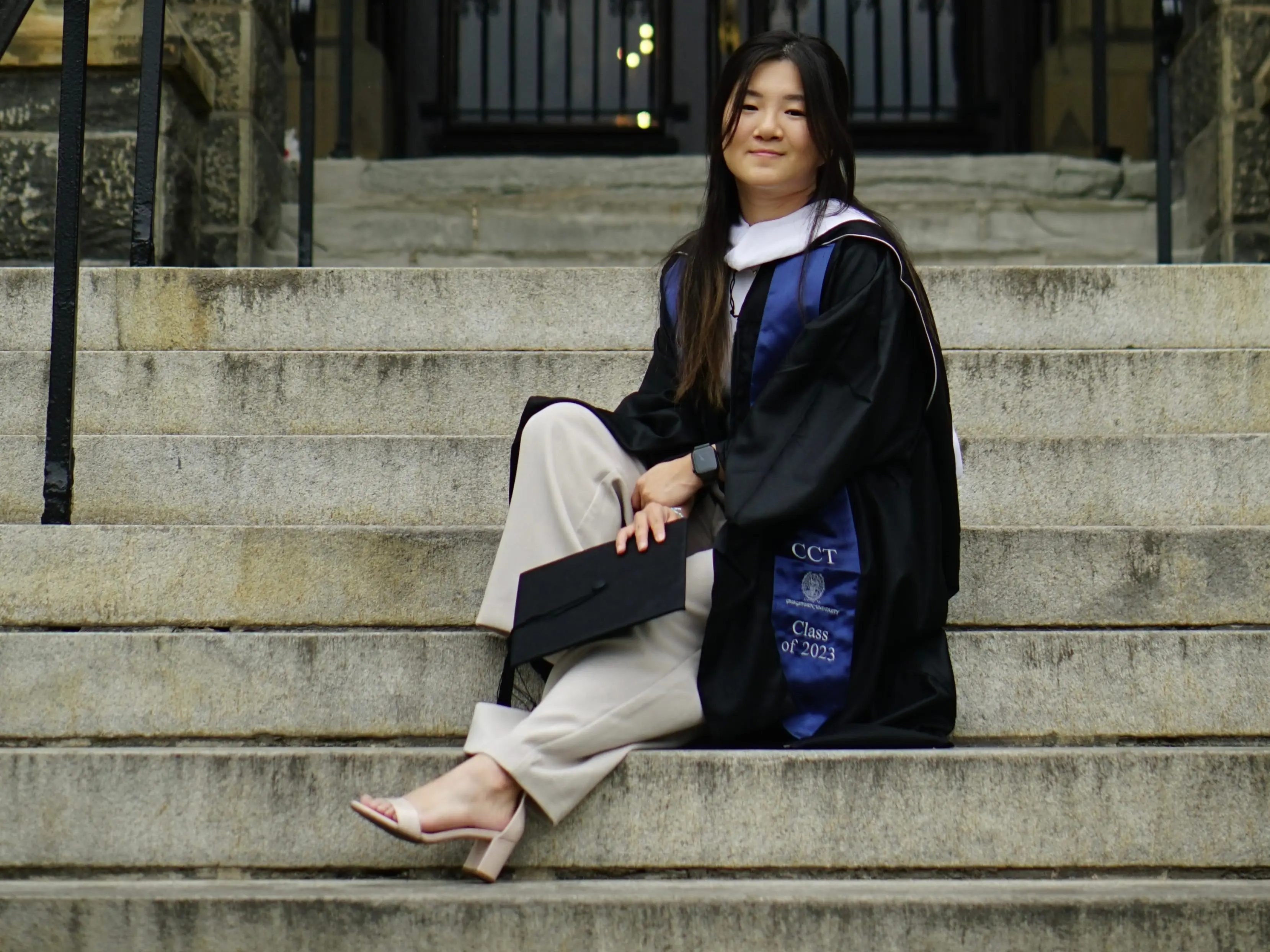 Graduate in capless academic regalia sits on stone steps outside an ornate arched building entrance.