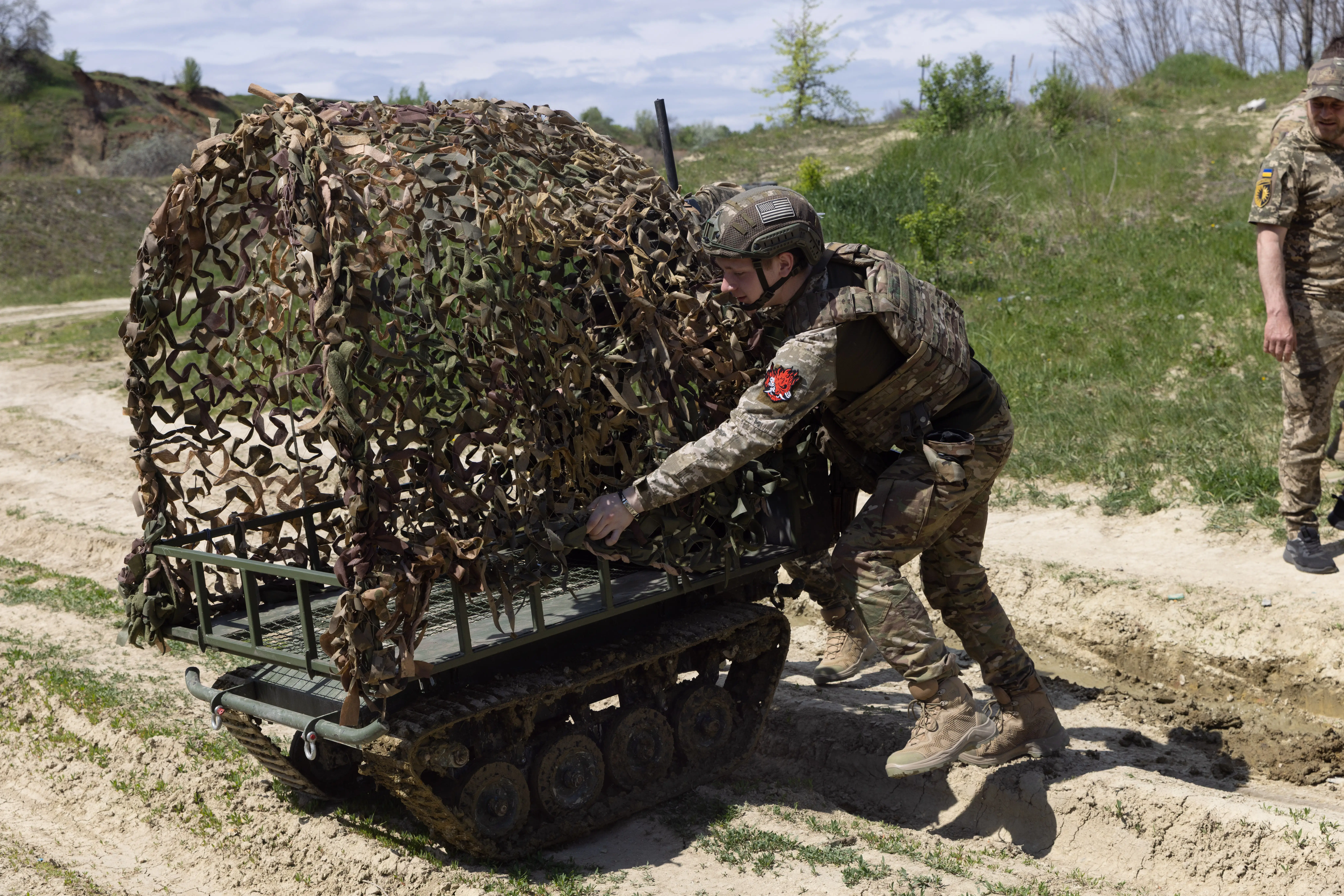 Uniformed personnel adjust camouflage netting on a small tracked ground vehicle along a dirt path.