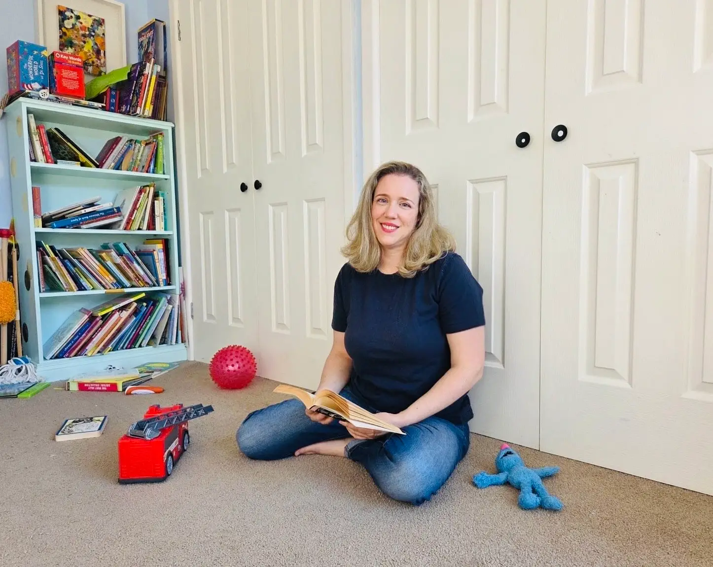 The author sits on the floor of a kid's room with an open book in her hand.