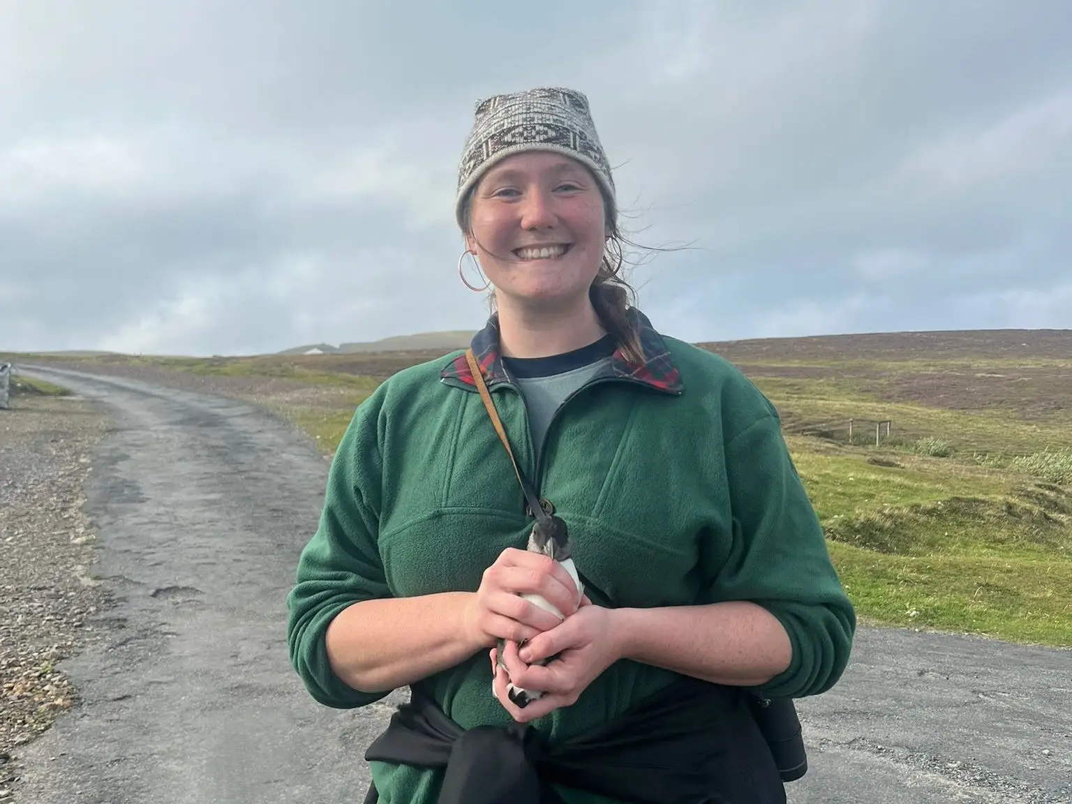The writer holds a small seabird on a rural road under a cloudy sky, on a remote island.