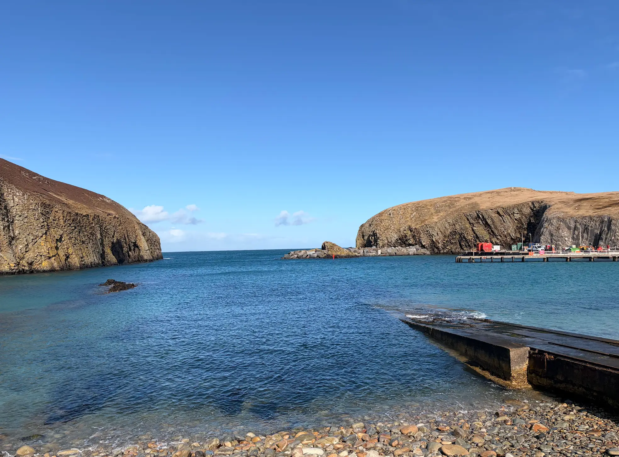 Rocky coastal harbor with blue water, cliffs, a pier, and a concrete slipway under a clear sky.