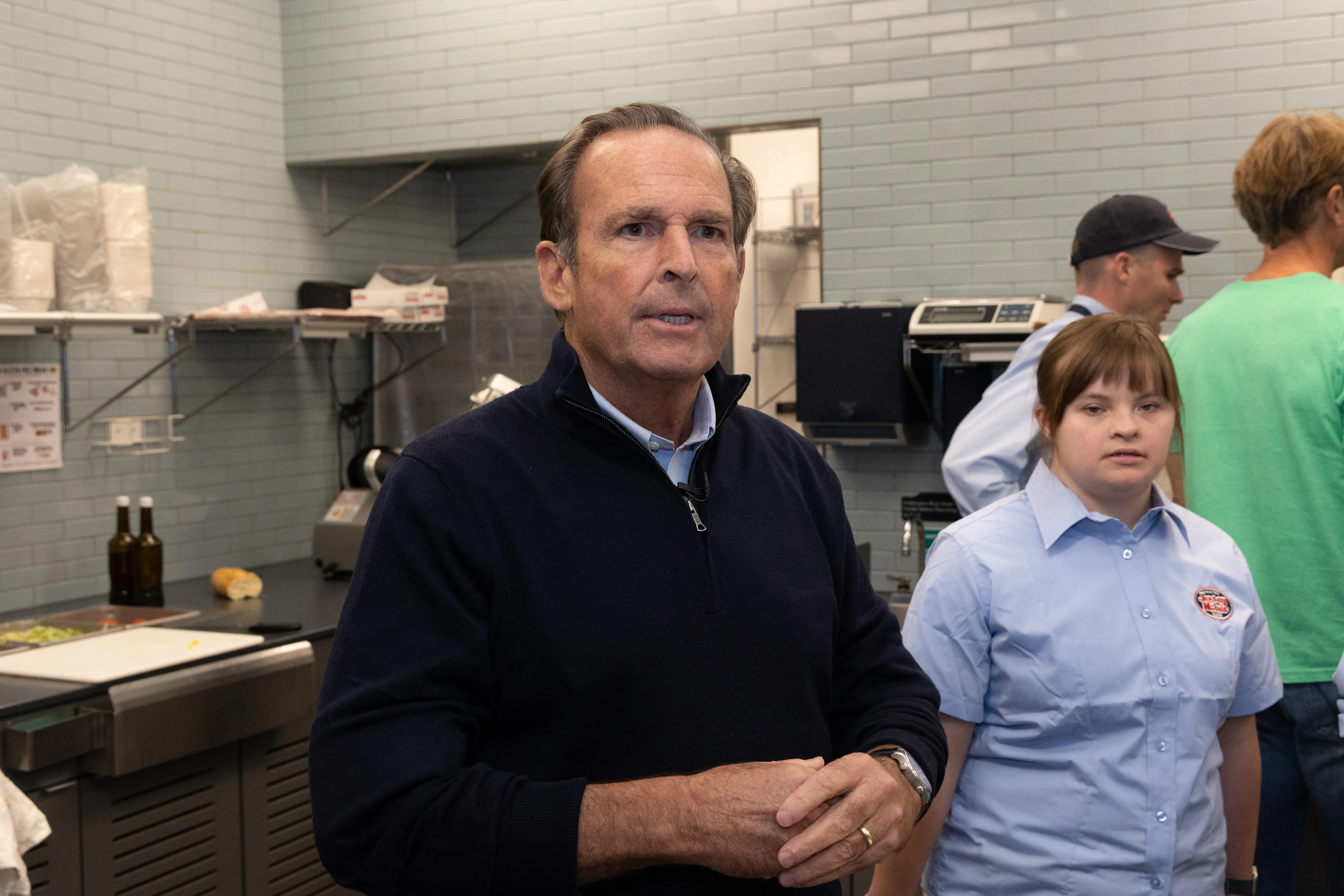 Jersey Mike's founder Peter Cancro stands in a kitchen while wearing a dark blue quarter-zip and a light blue dress shirt underneath.