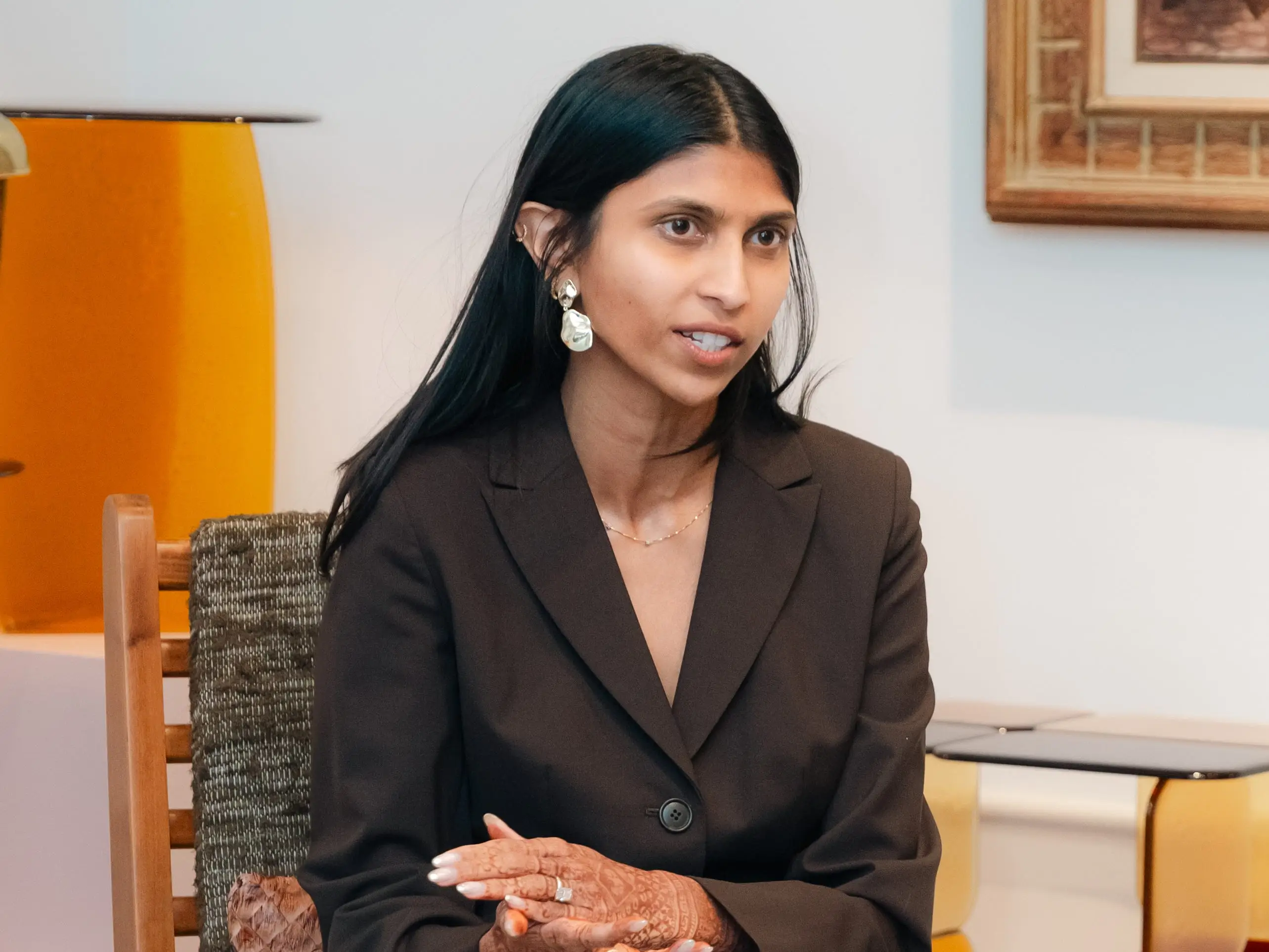 Maya Bakhai sits on a chair with a framed piece of artwork on the wall behind her.