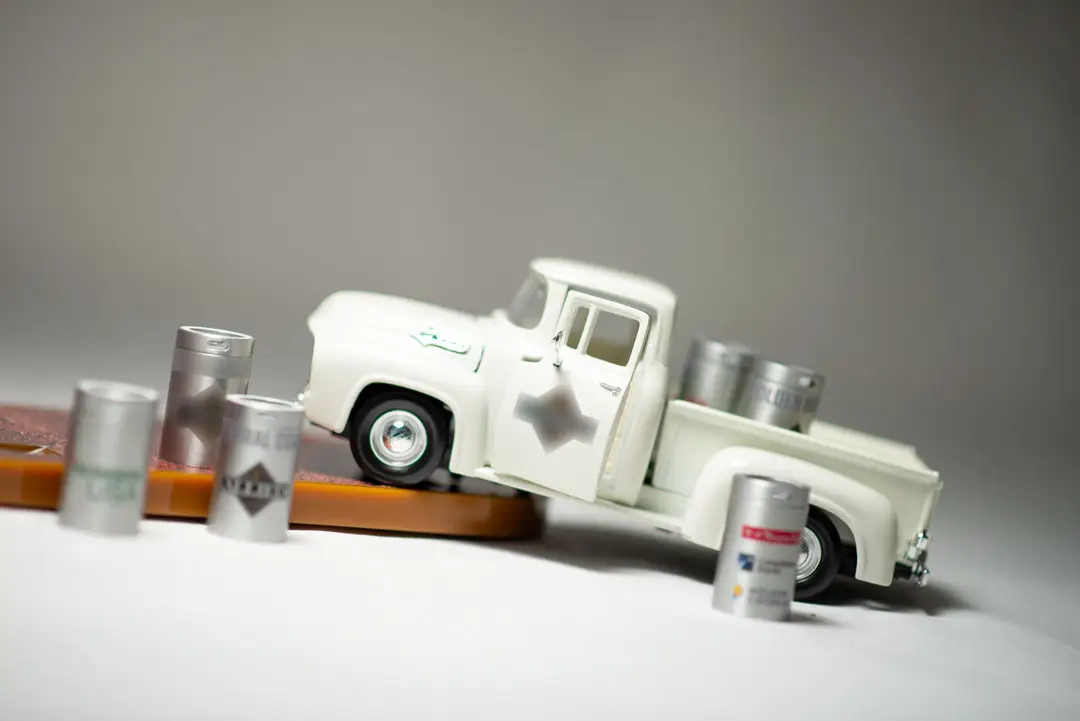 Miniature white pickup truck sits among small silver barrels on a neutral studio surface.