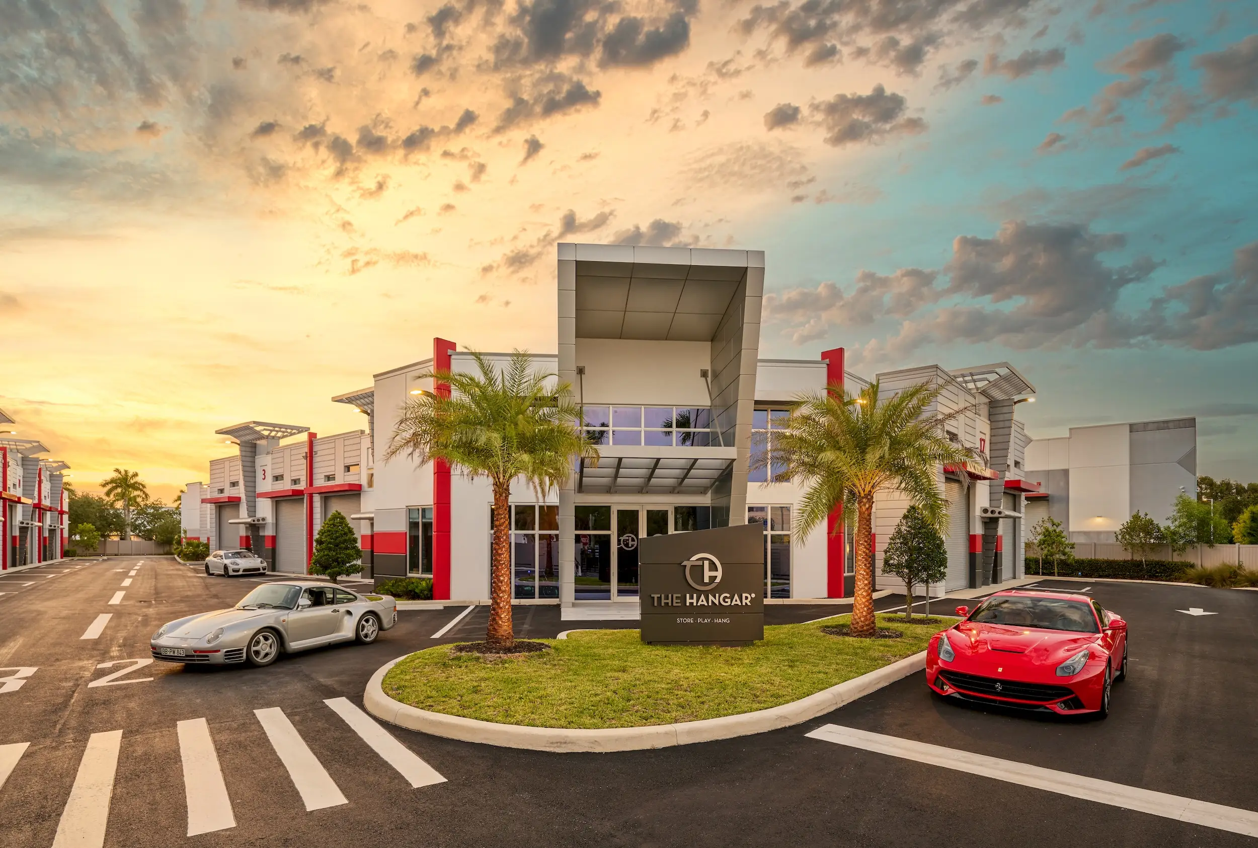 The exterior of a car storage facility with palm trees and sports cars parked outside at sunset.