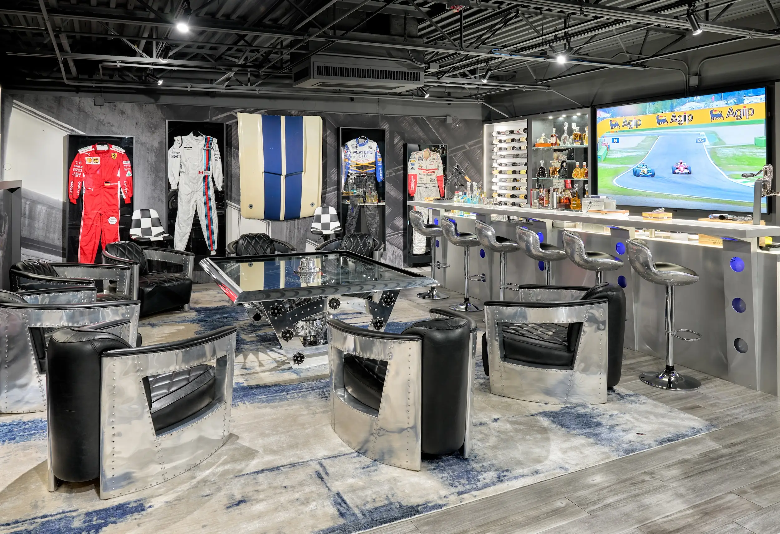 Motorsport-themed lounge with racing suits, polished metal furniture, bar seating, and a television showing a race broadcast.