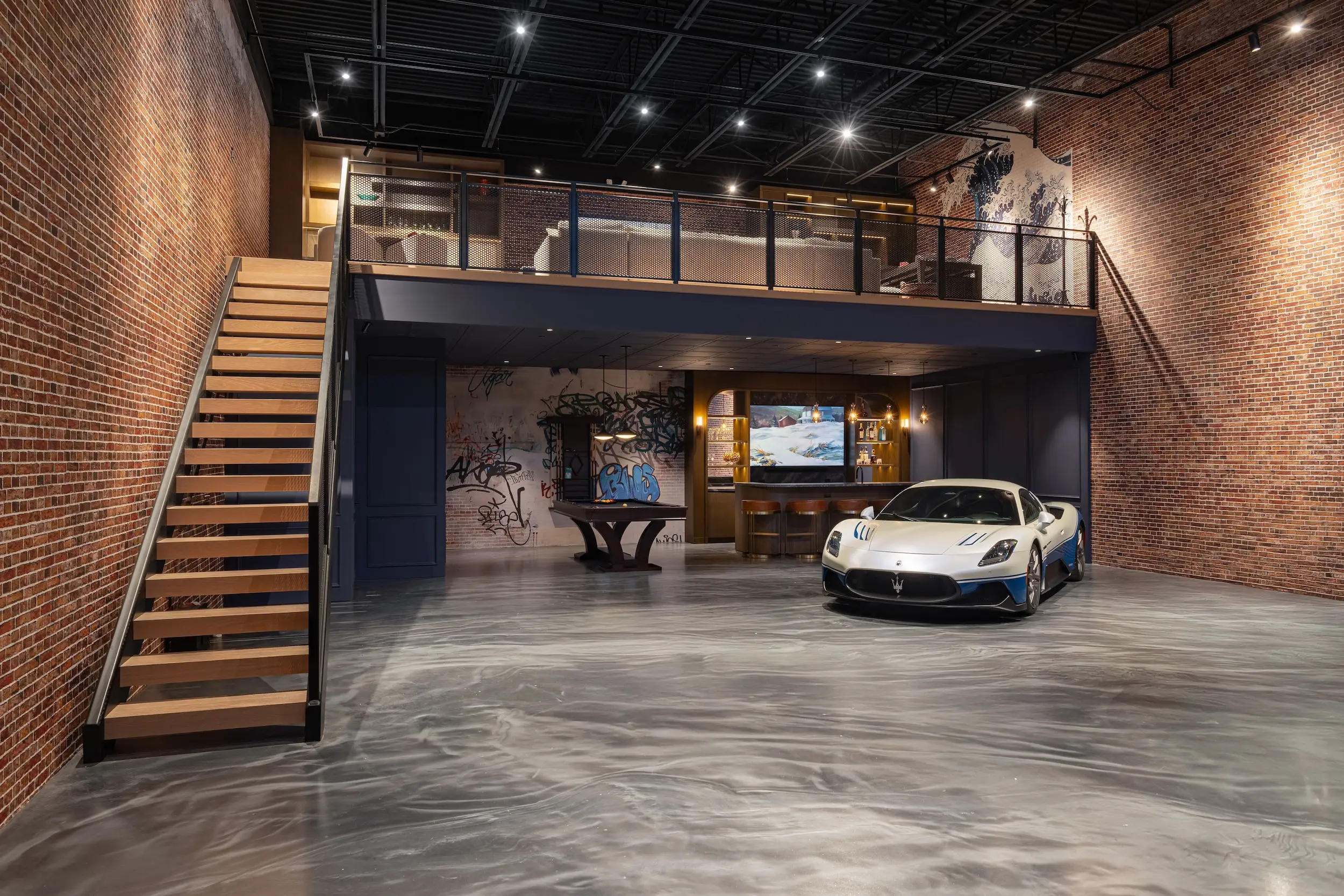 White Maserati sports car parked inside a brick-walled loft garage with a staircase, mezzanine, and bar area.