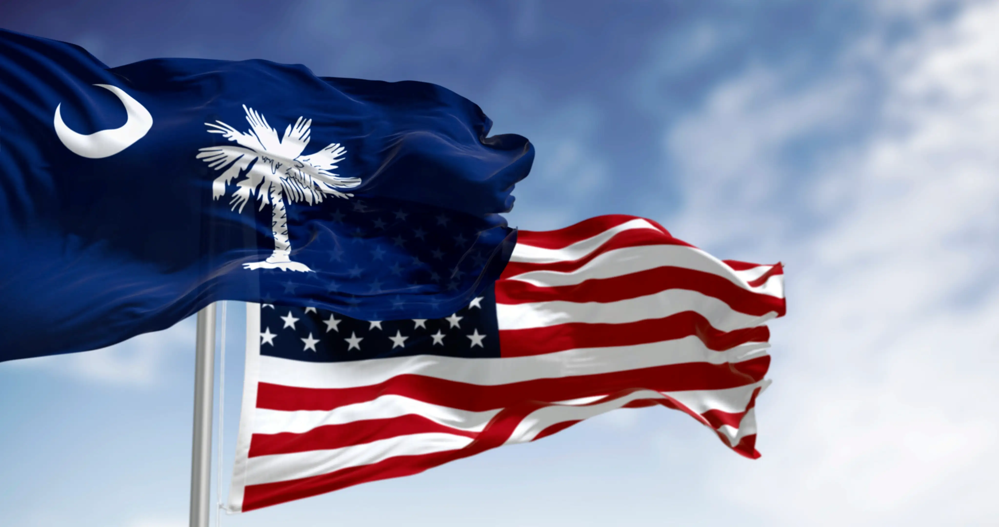 South Carolina state flag and United States flag wave together against a cloudy blue sky.