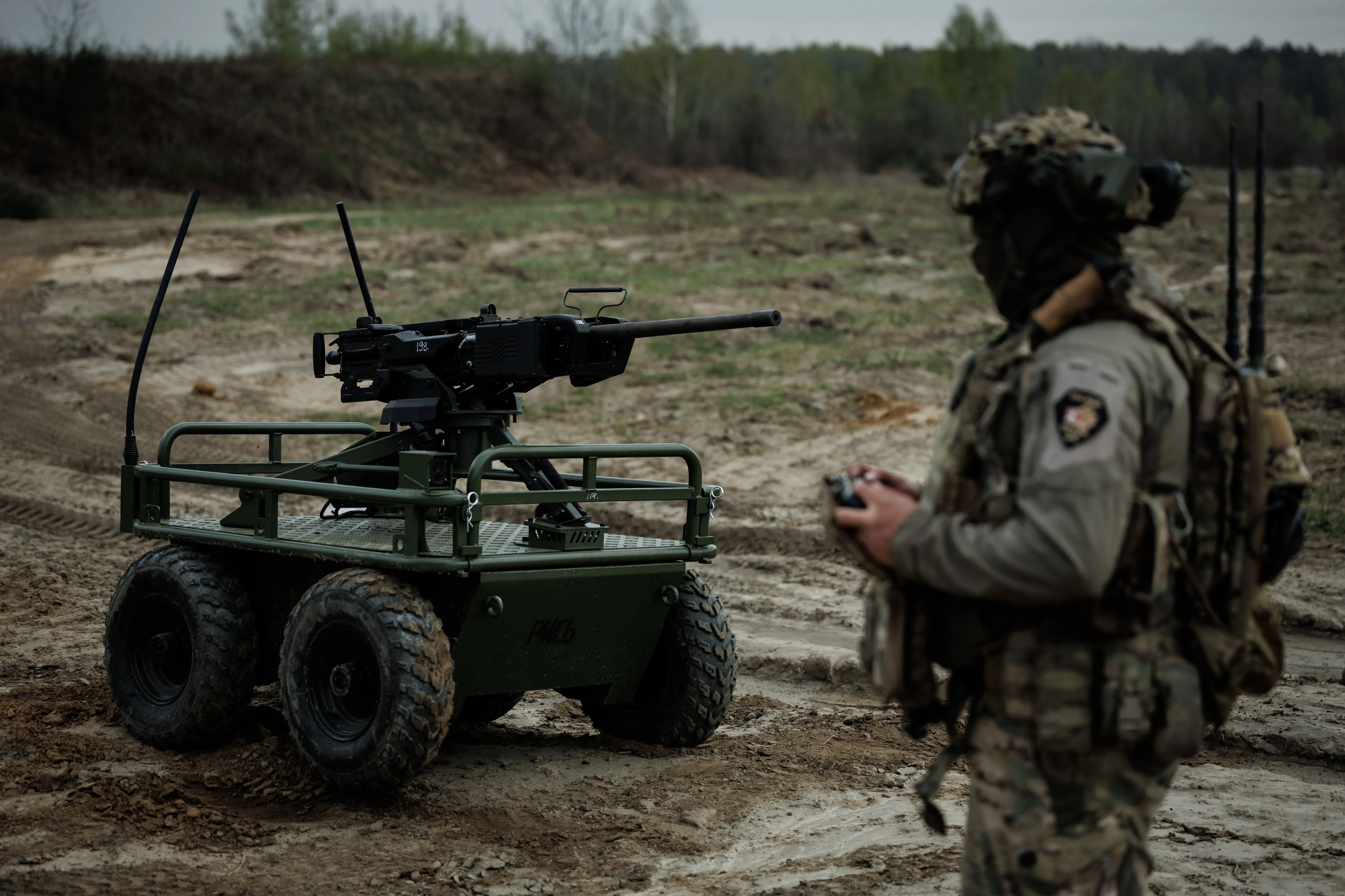 Camouflaged service member operates a four-wheeled unmanned ground vehicle with a mounted weapon on a muddy range.