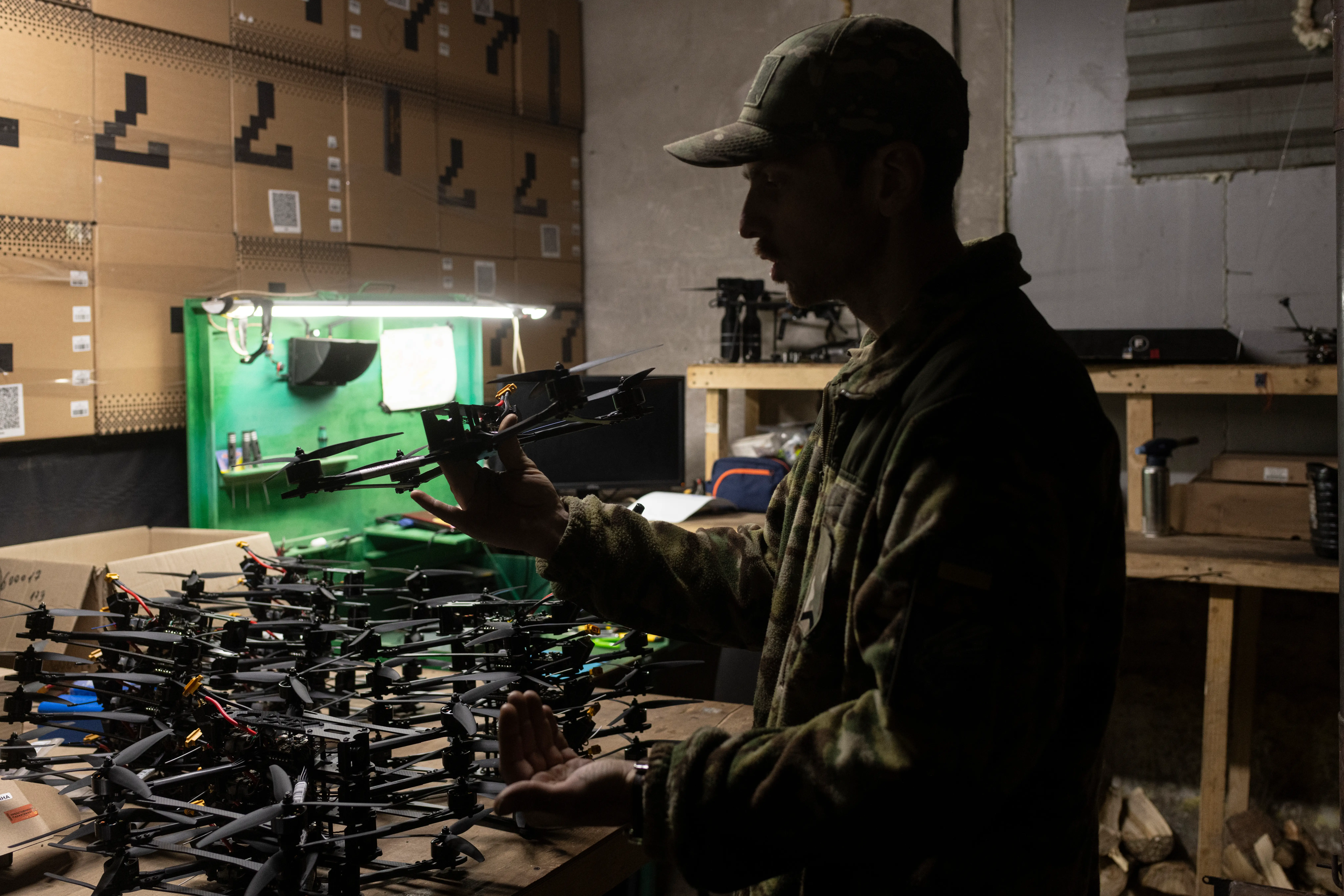 Person in a cap inspects small quadcopter drones on a cluttered workshop bench under dim lighting.