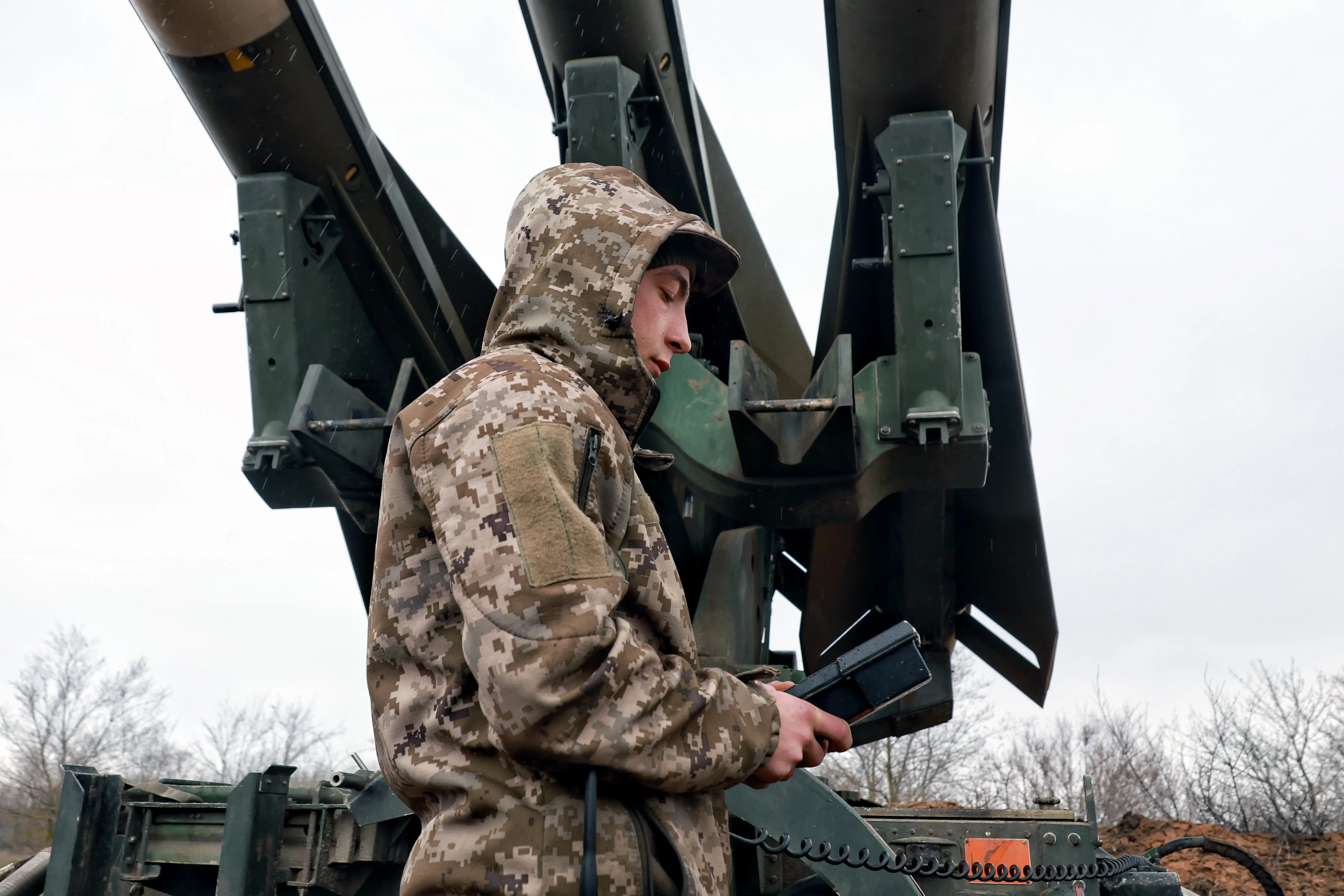 A person in camouflage uniform operates a handheld device beside large mounted military launcher tubes outdoors.