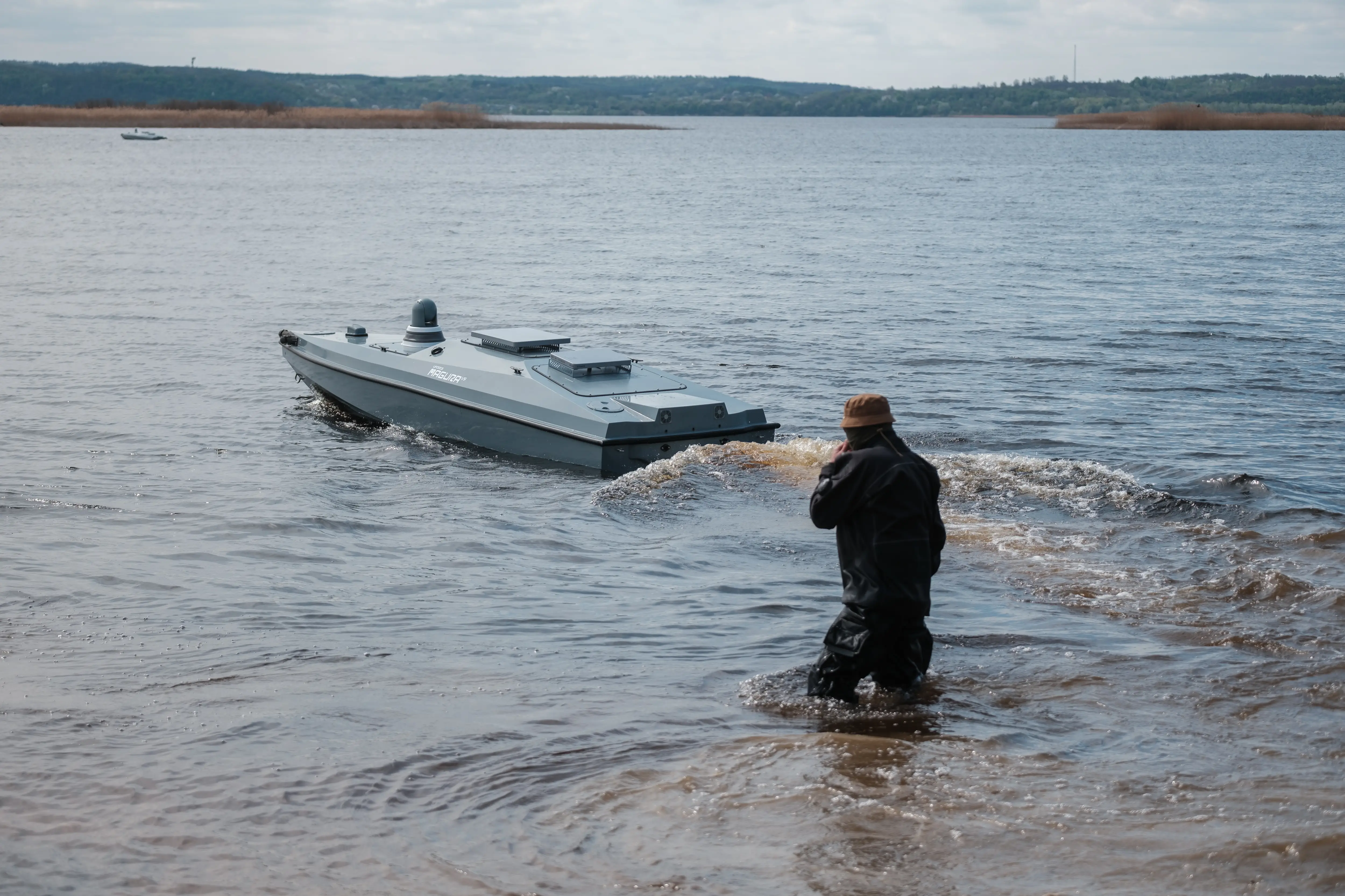 A person stands in shallow water near a small unmanned-looking boat moving across a wide river or lake.
