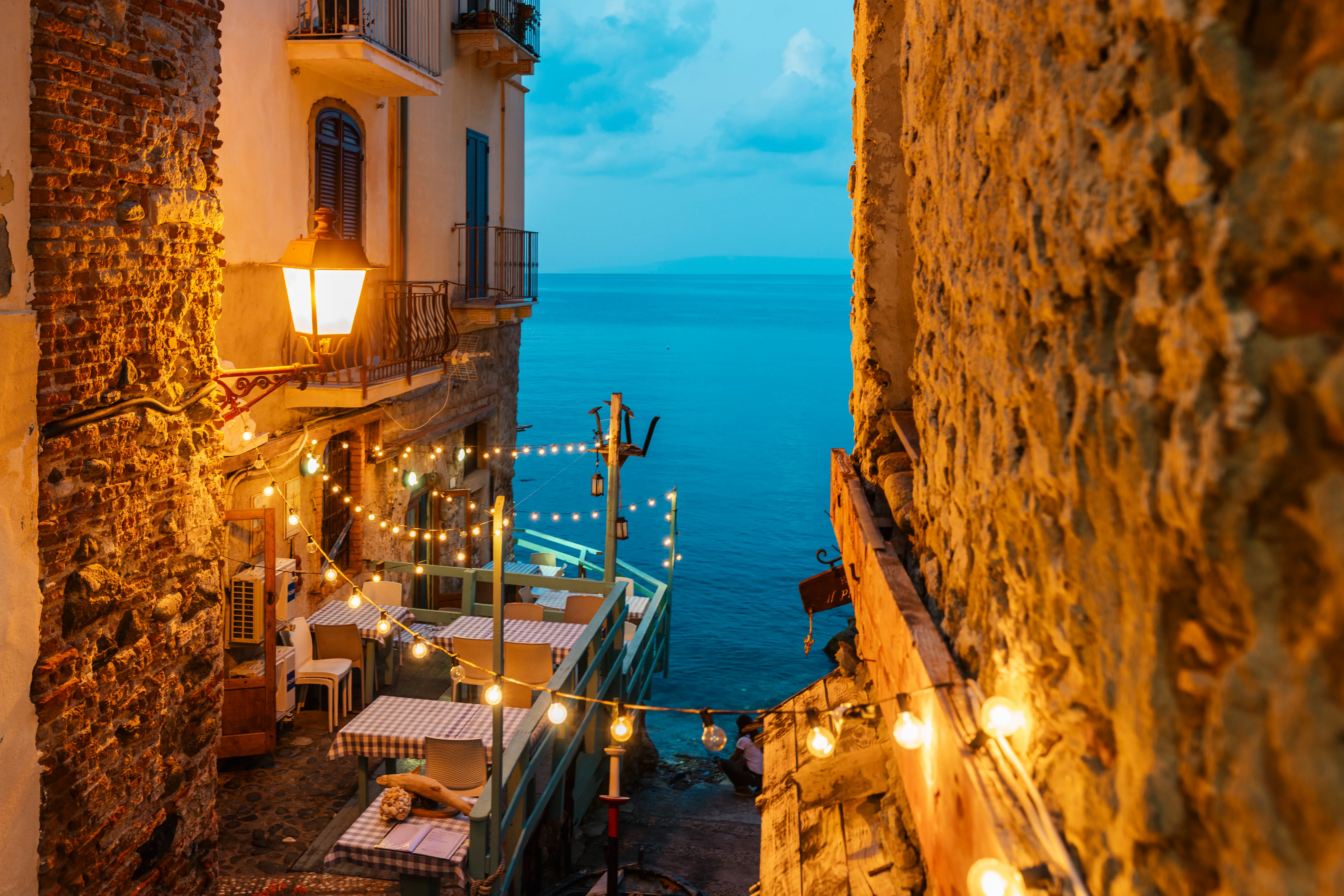Warm string lights and a lantern illuminate a seaside restaurant terrace glimpsed between stone walls at blue hour, the Chianalea district of Scilla, Calabria, Italy.