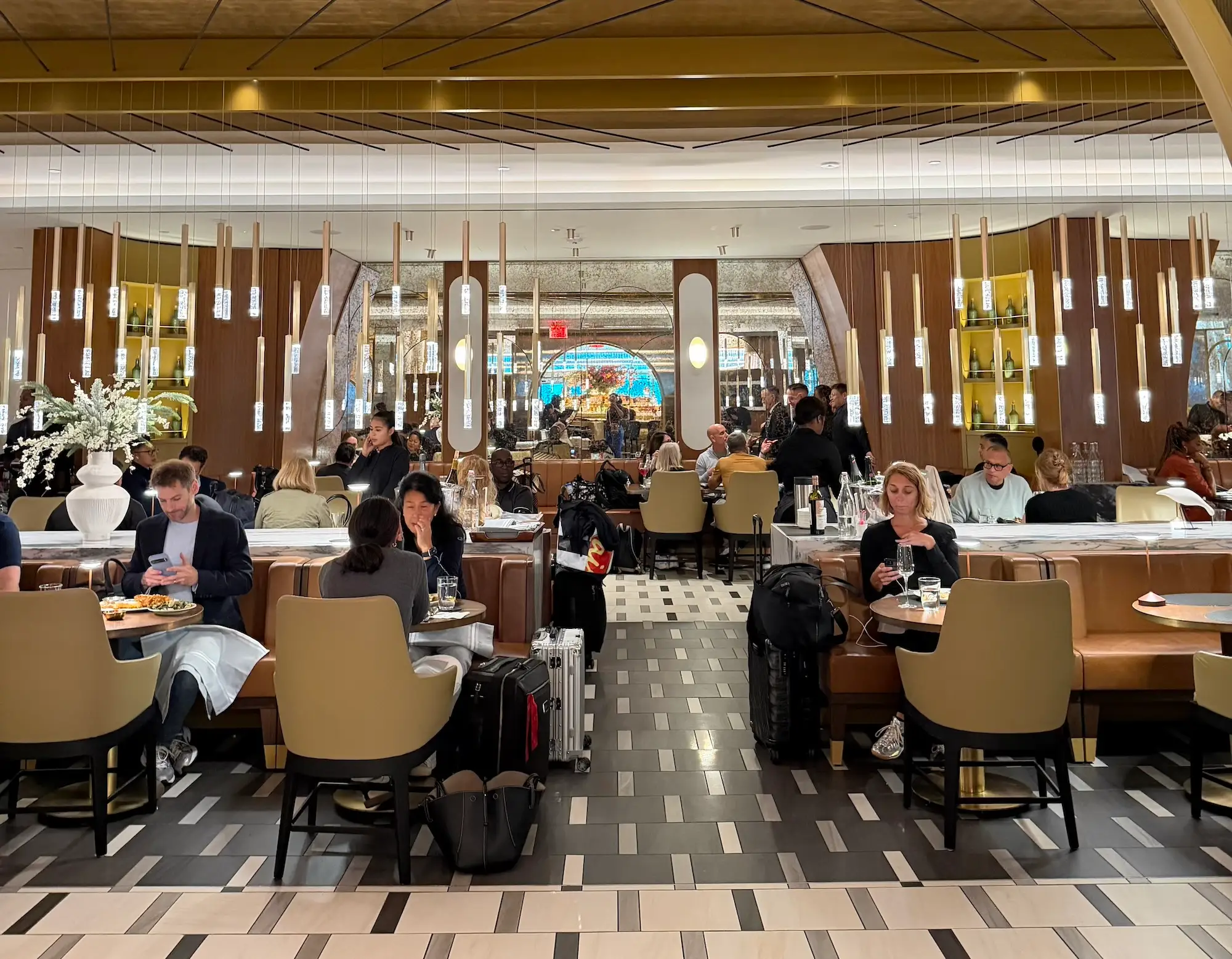 Busy modern restaurant dining area with seated patrons, luggage, pendant lights, booths, and a reflective decorative interior.