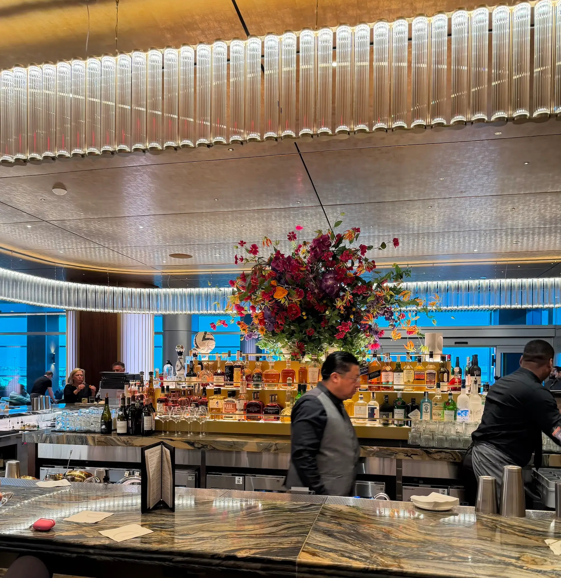 Bartenders work behind an illuminated bar with liquor bottles and a large floral arrangement.