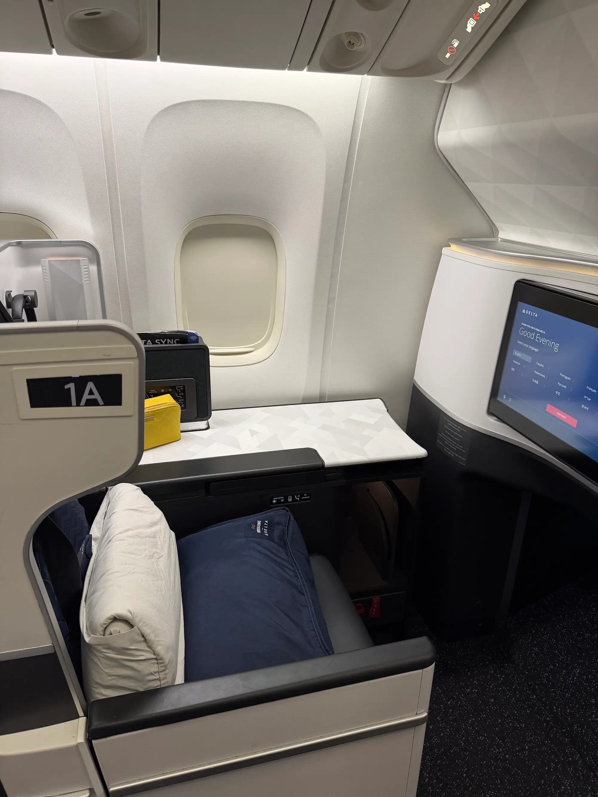 Business-class airplane seat 1A with pillows, window, side table, overhead controls, and seatback entertainment screen.