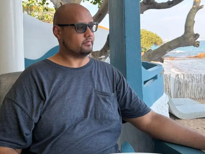A person wearing sunglasses sits at a seaside café table with trees and water visible behind them.