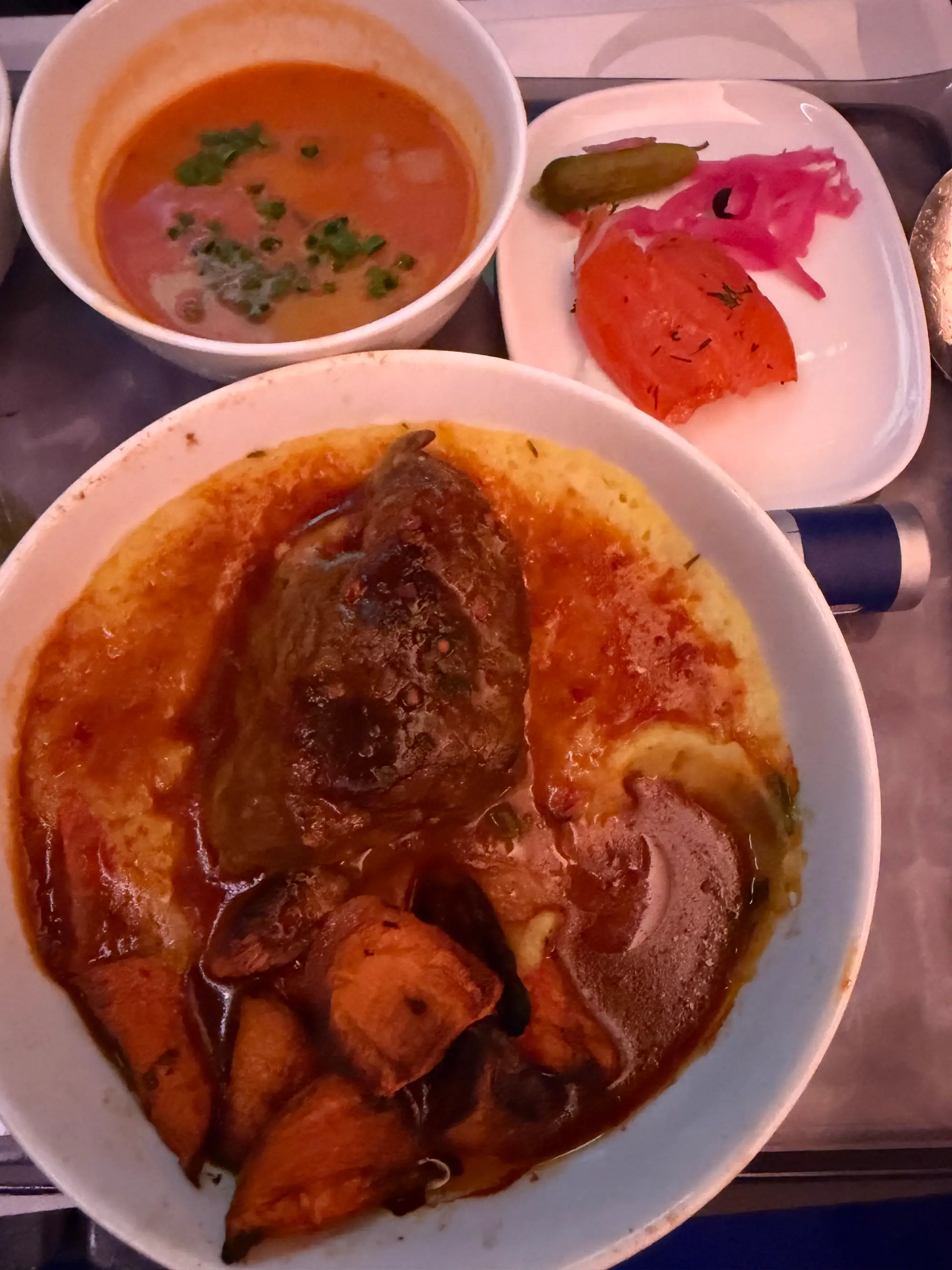 Tray with a bowl of sauced vegetables, a cup of orange soup, and a small plate of pickles and tomato.