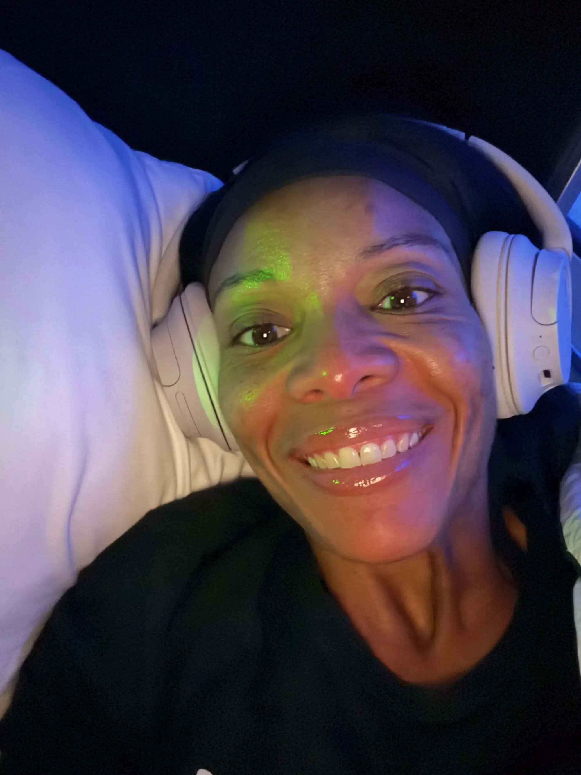 Smiling person wearing white over-ear headphones lies on a pillow under colorful blue and green lighting.