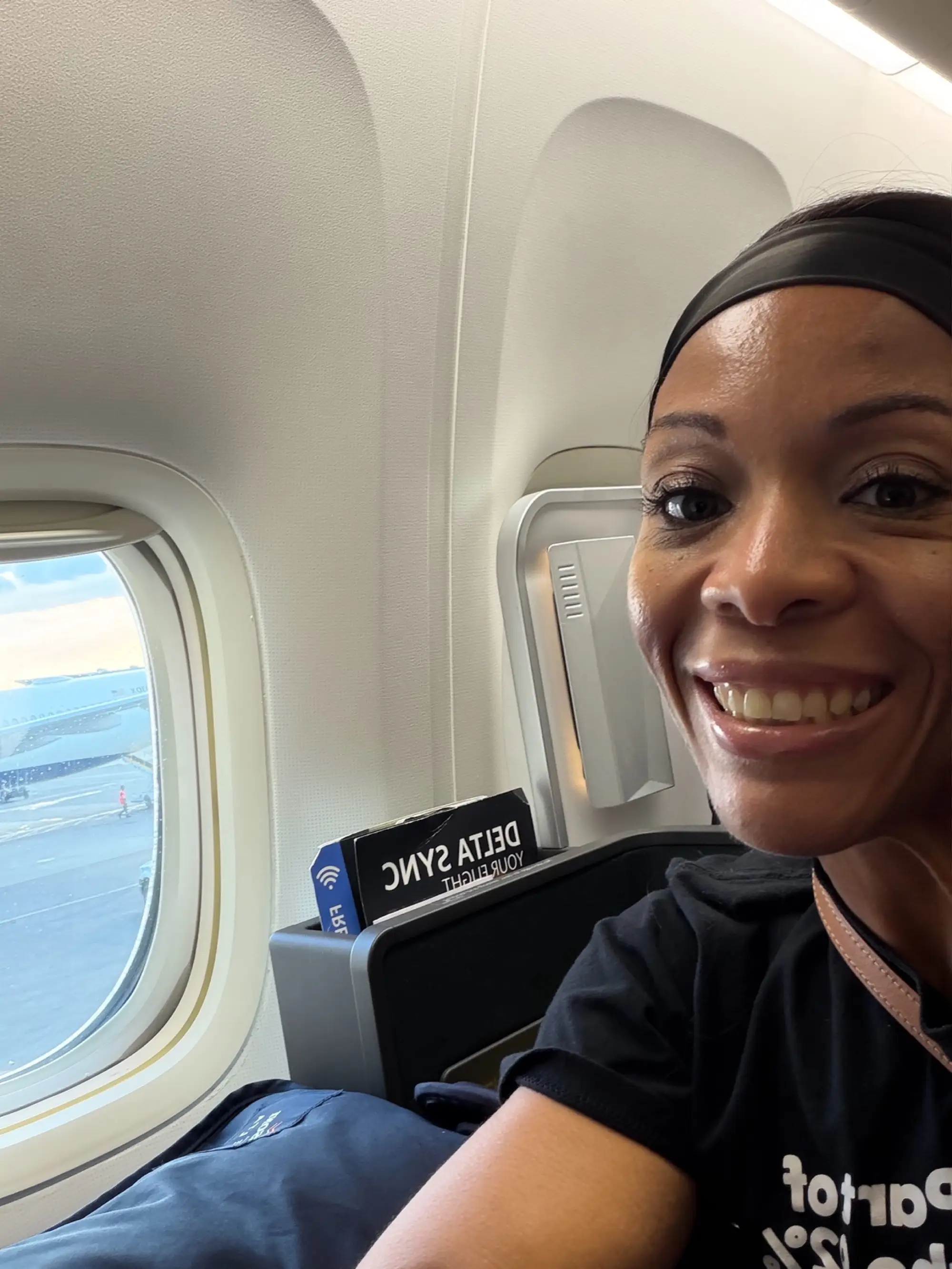 Smiling passenger seated by an airplane window with a Delta Sync seatback sign visible.