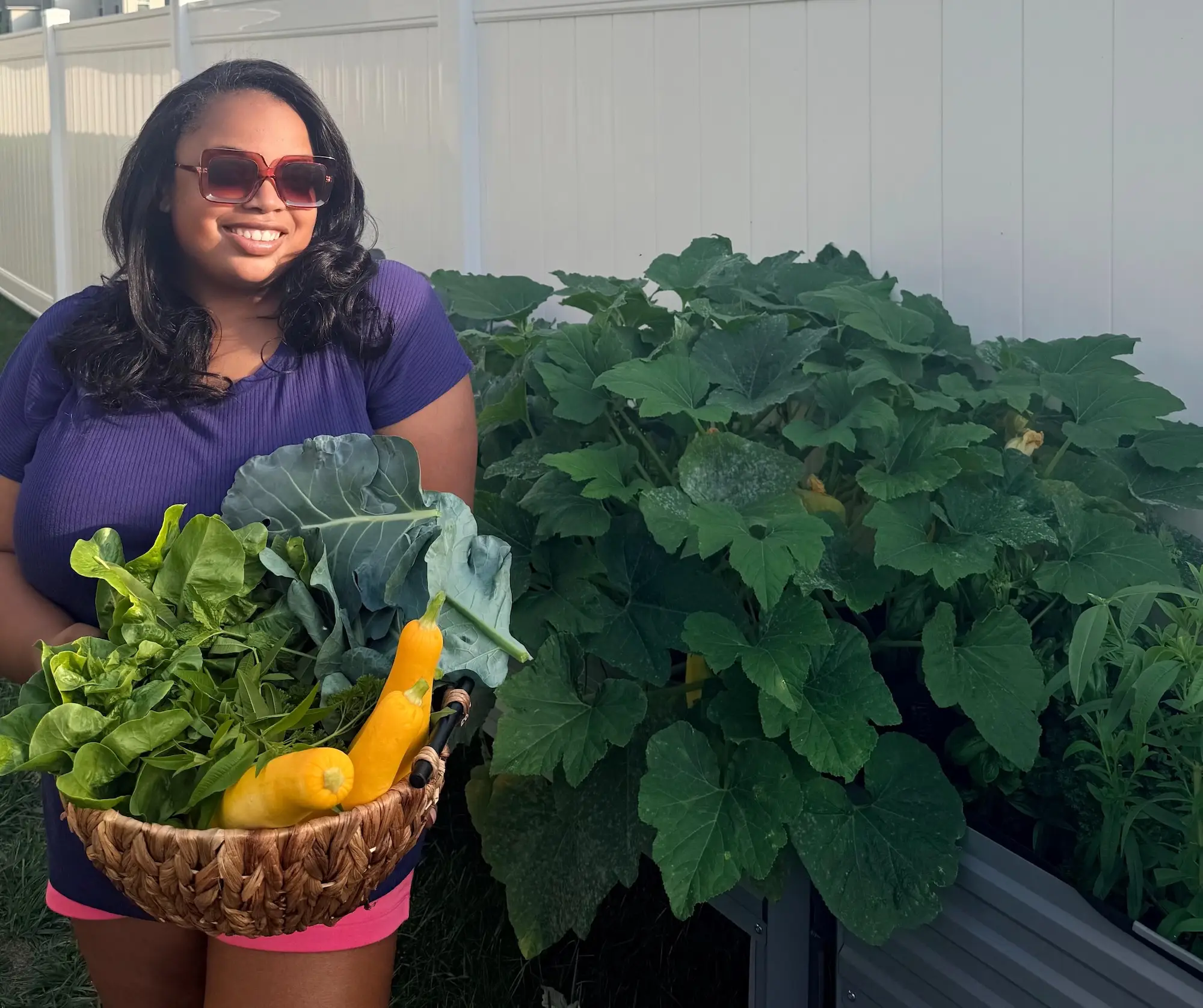 Smiling person in sunglasses holds a basket of leafy greens and yellow squash beside a lush raised garden bed.