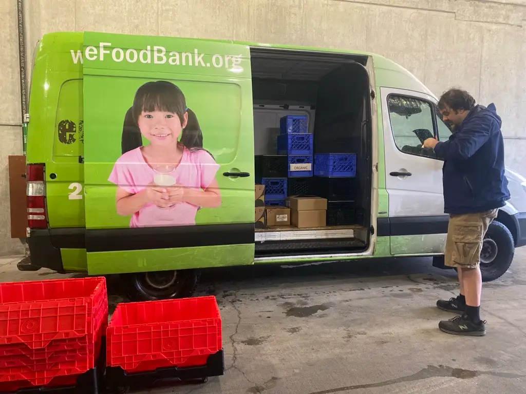 A West Seattle Food Bank employee stands at the right of the frame in front of a green food bank can. Black-and-red plastic totes stand in the foreground.