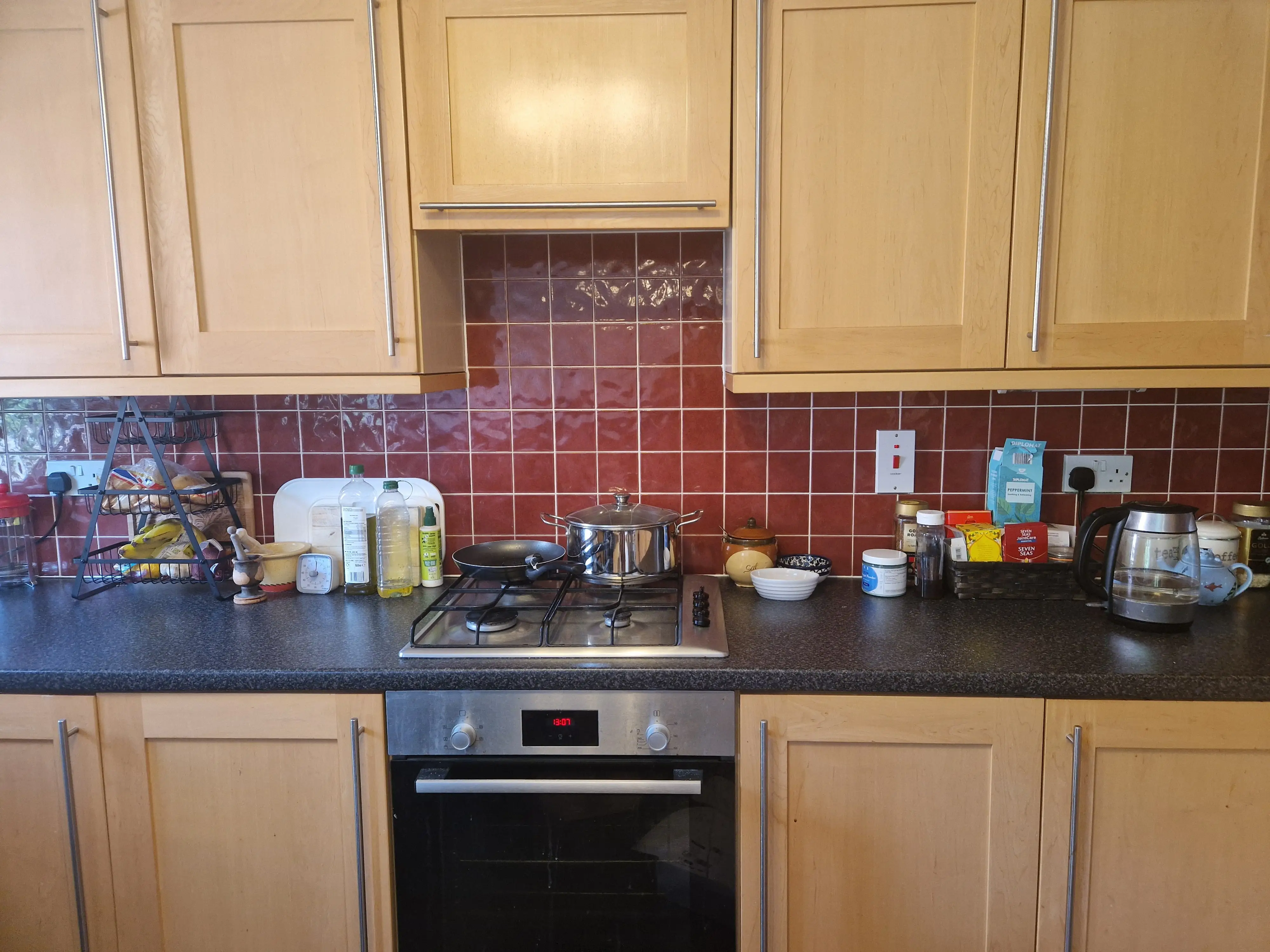 The writer's apartment kitchen that features wooden cabinets, a red tile backsplash, an oven, a stovetop, and a variety of kitchenware and groceries.