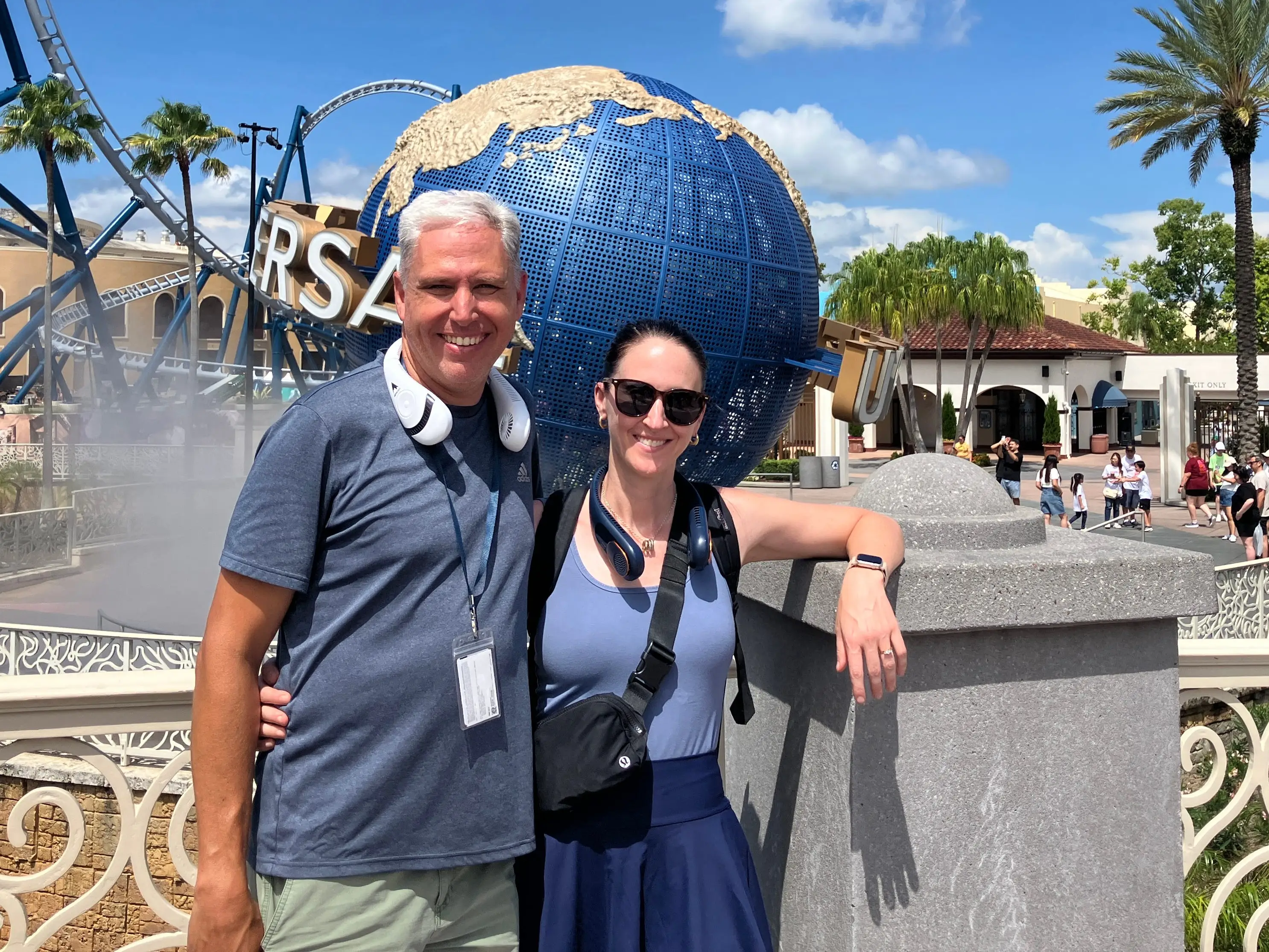Two visitors pose in front of the Universal globe fountain and roller coaster at a sunny theme park.
