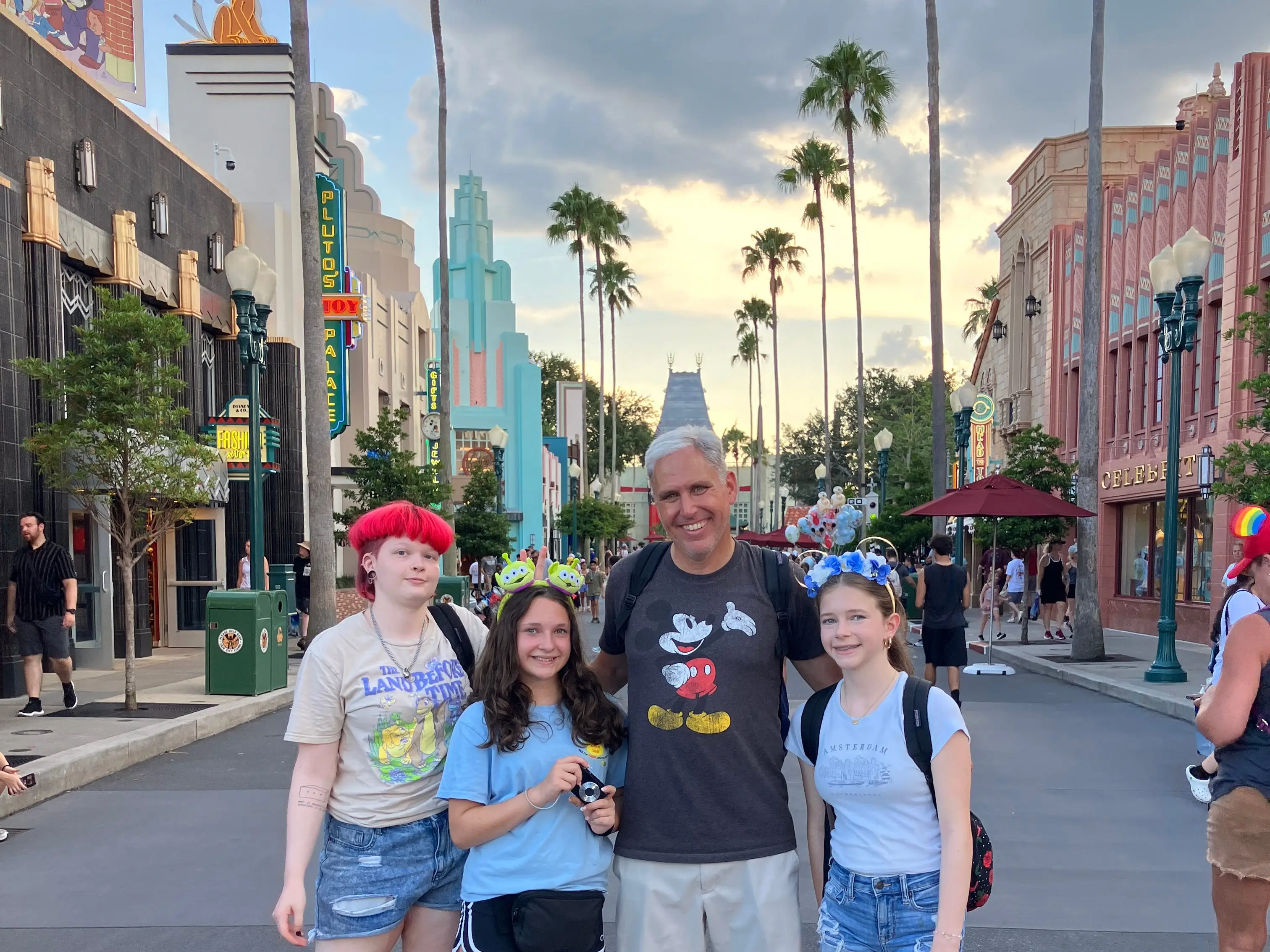 Four park visitors pose on a palm-lined Disney street with Art Deco storefronts and a stormy evening sky.