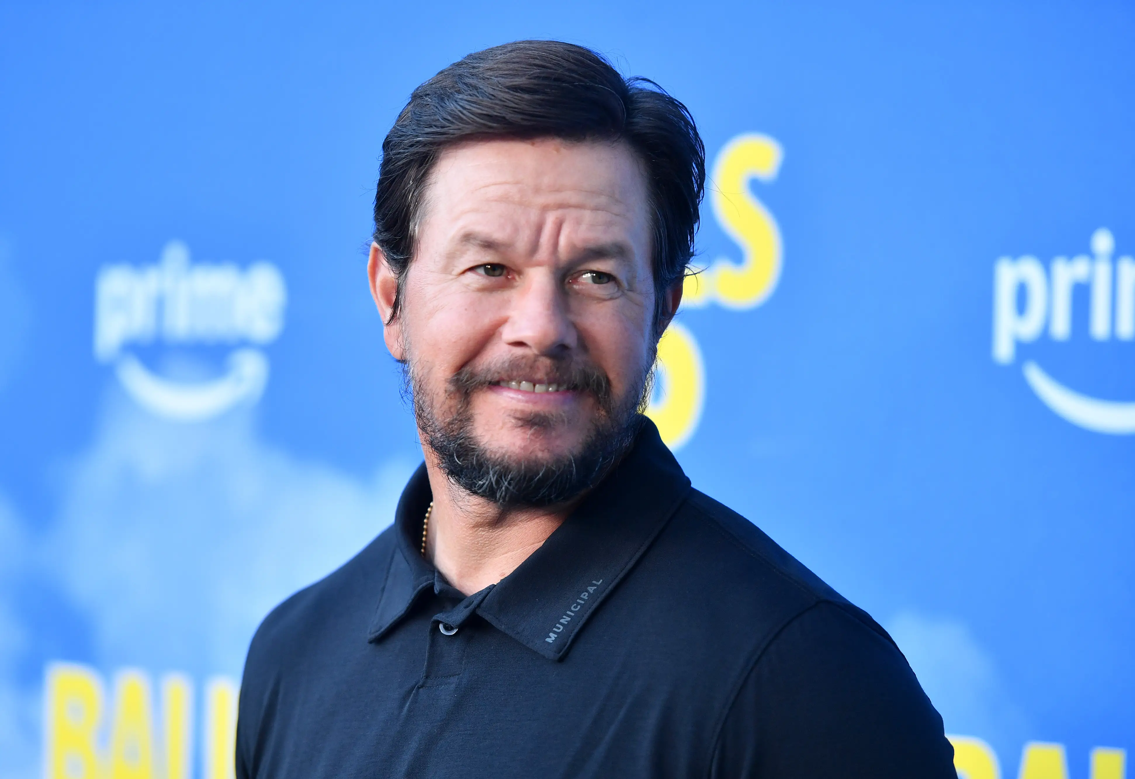 Mark Wahlberg poses on a blue Amazon Prime event backdrop with yellow promotional lettering.